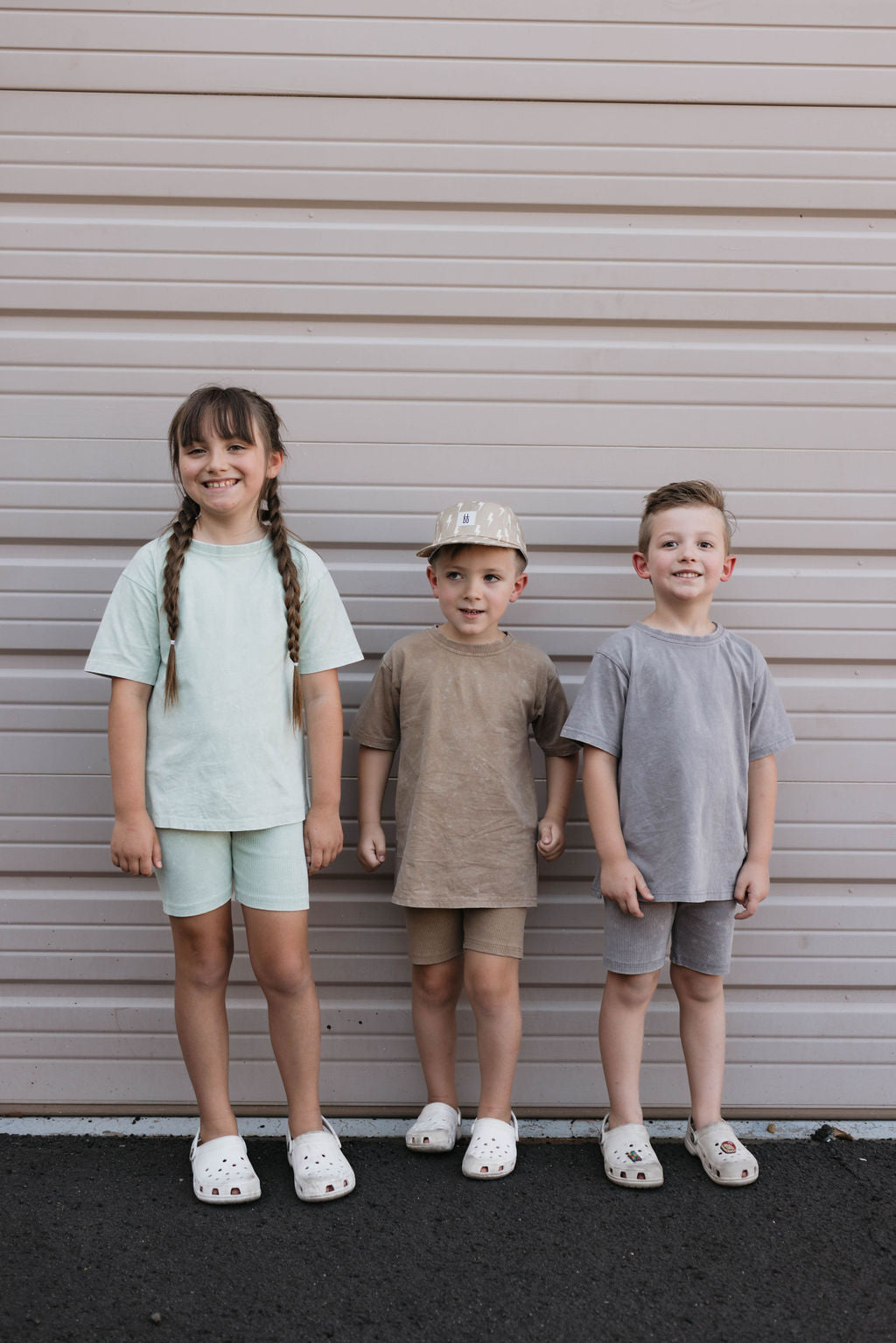 Three children stand in front of a beige wall, wearing adorable outfits from forever french baby. The girl on the left has long brown hair in braids and wears a light green outfit made of organic cotton. The middle boy sports the Children's Short Set in Vintage Washed Steel along with a matching hat, while the boy on the right wears grey. All three are smiling in their white Crocs.