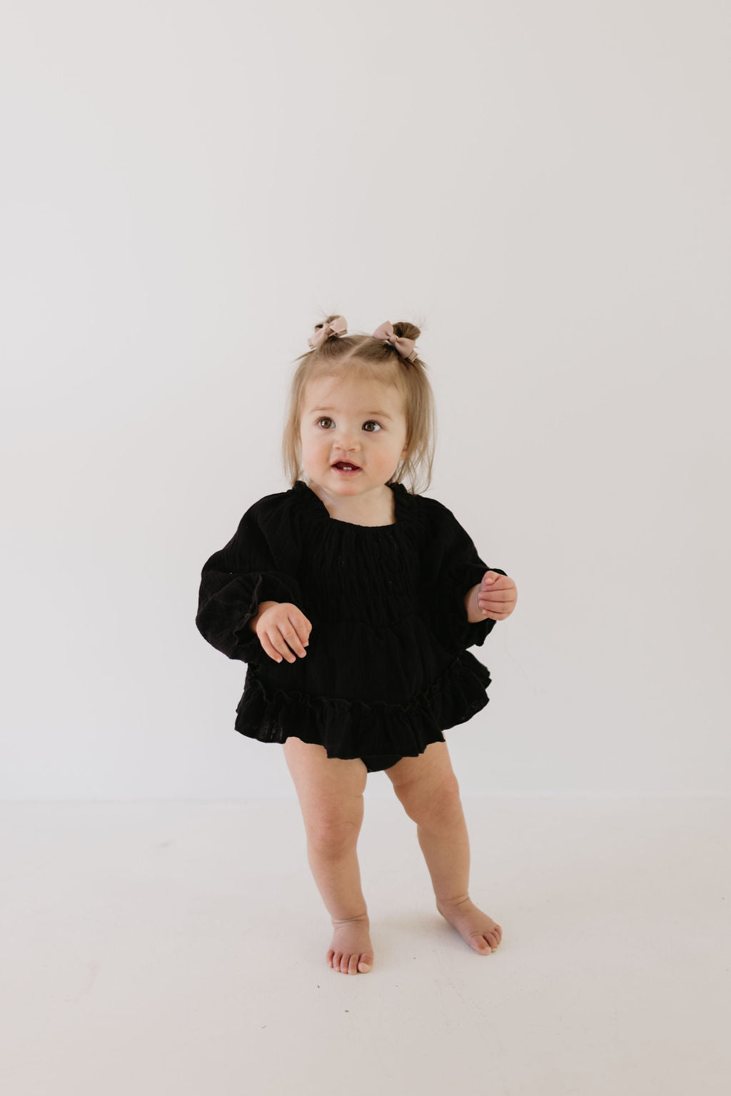 A baby is barefoot against a plain white backdrop, dressed in a black "Ruffle Romper | Aria" by forever french baby. Her hair, styled in small pigtails with bows, adds charm as she gazes slightly to her left with a curious expression.