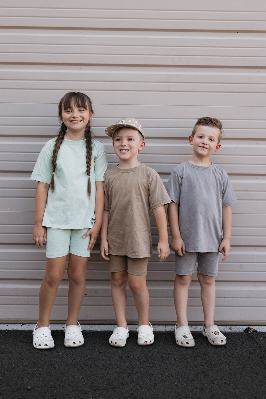 Three children stand side by side in front of a gray garage door. The child on the left wears Forever French Baby's Vintage Washed Spearmint Children's Short Set with braids and white shoes. The middle child sports a vintage washed beige outfit with a cap and white shoes, while the child on the right dons a gray oversized tee and ribbed shorts paired with white shoes.