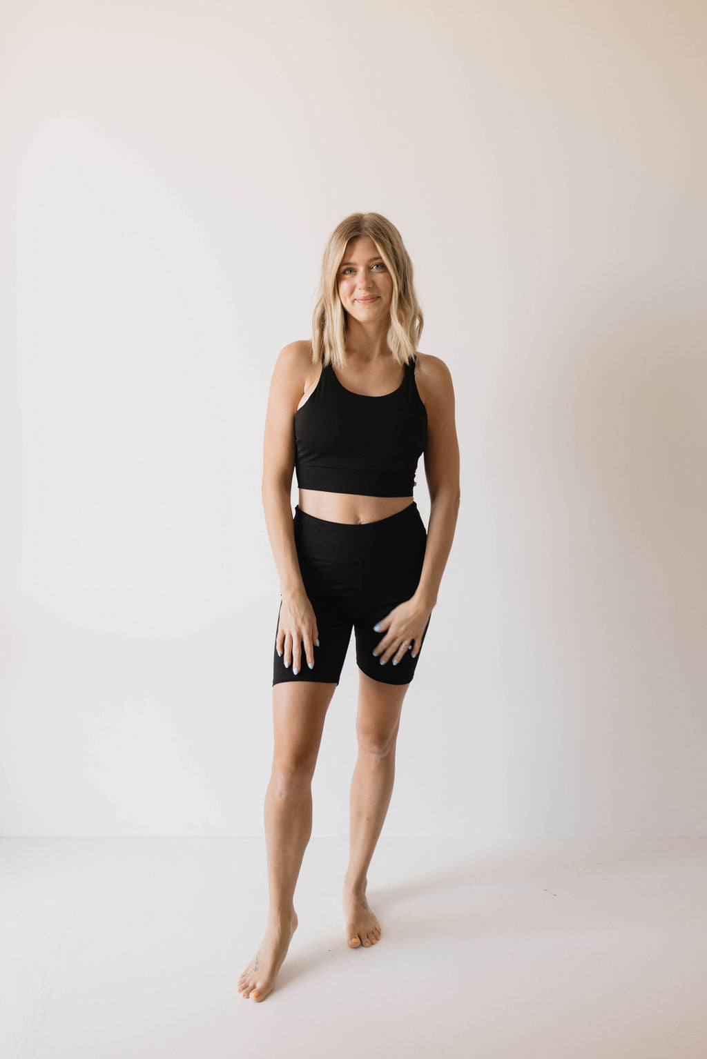 A woman stands against a plain white background wearing the Women's Workout Set in black by forever french baby, consisting of a sports tank top and biker shorts. She has blonde shoulder-length hair and is barefoot. Smiling gently, her hands rest on her thighs.