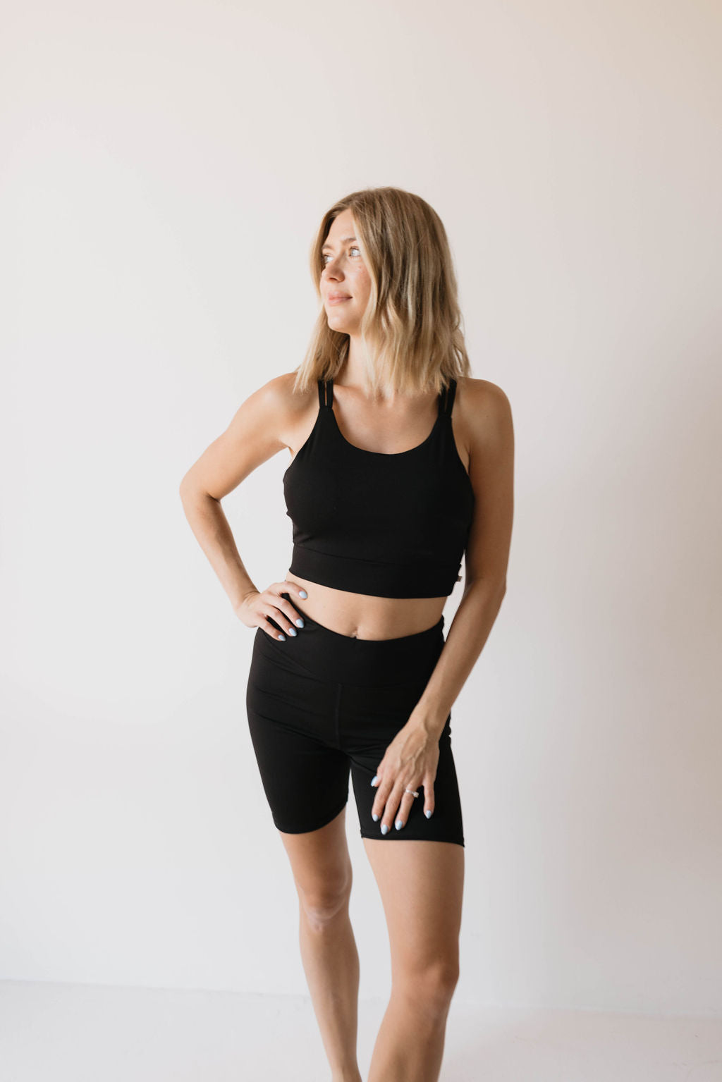 A woman with shoulder-length blonde hair is standing against a plain white background, wearing a stylish Women's Workout Set in Black by forever french baby. One hand rests on her hip while the other is placed on her thigh, as she gazes slightly to the side.