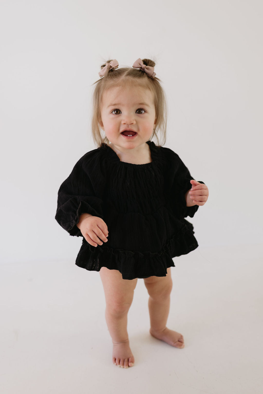 A toddler with light brown hair, adorned with bows, wears a loose black "Ruffle Romper | Aria" by forever french baby. She stands barefoot against a plain white background, smiling at the camera.