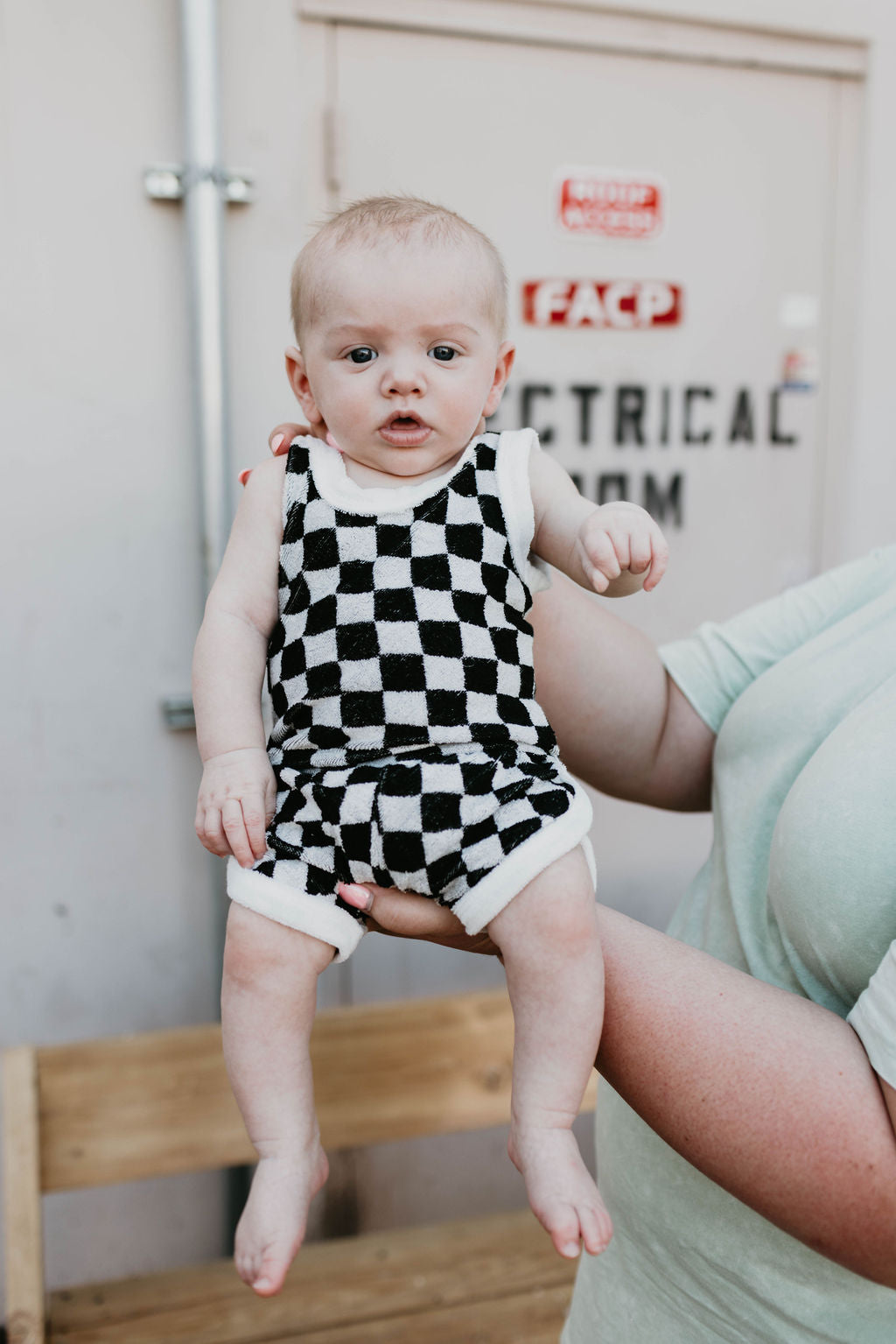 A baby wearing a stylish Terry Tank Short Set in Black + White Checker by Forever French is being supported by an adult's hand. The background showcases a door with warning signs and visible pipes. The baby has a curious expression while the partially visible adult helps hold them up.