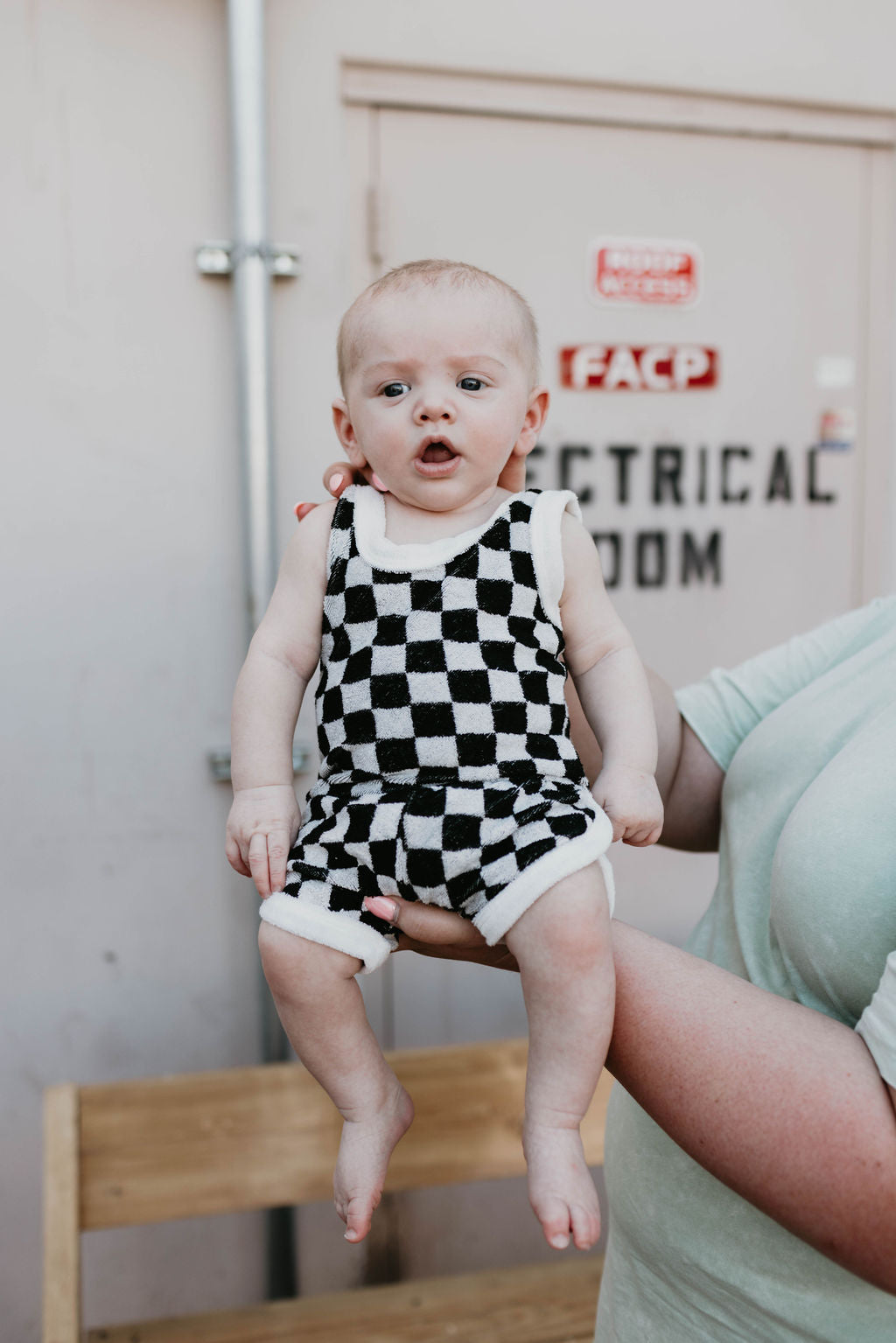 An adult, partially visible in a green shirt, is holding up a baby dressed in the trendy Forever French Terry Tank Short Set in black and white checker print. The backdrop features a beige wall with a sign that reads "Electrical Room.