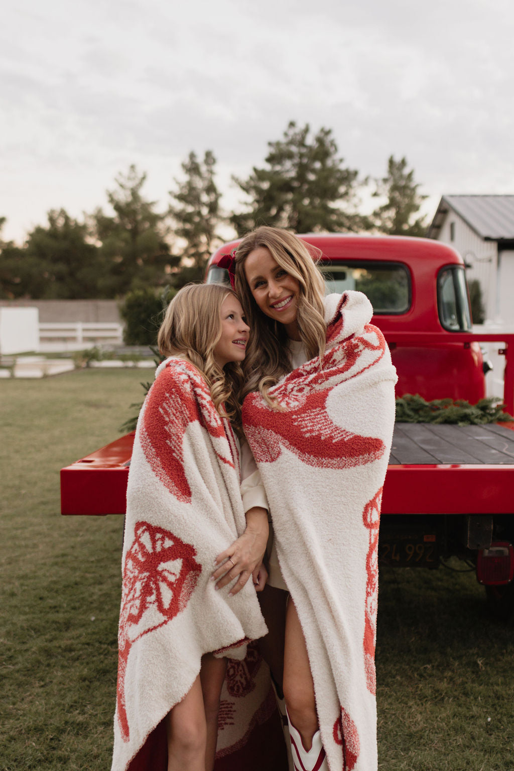 A mother and daughter wrapped in lolo webb's dreamy Plush Blanket | Cowgirl Christmas stand beside a red vintage truck. They are smiling in a grassy field with trees and a building in the background, creating a warm and cozy outdoor scene.