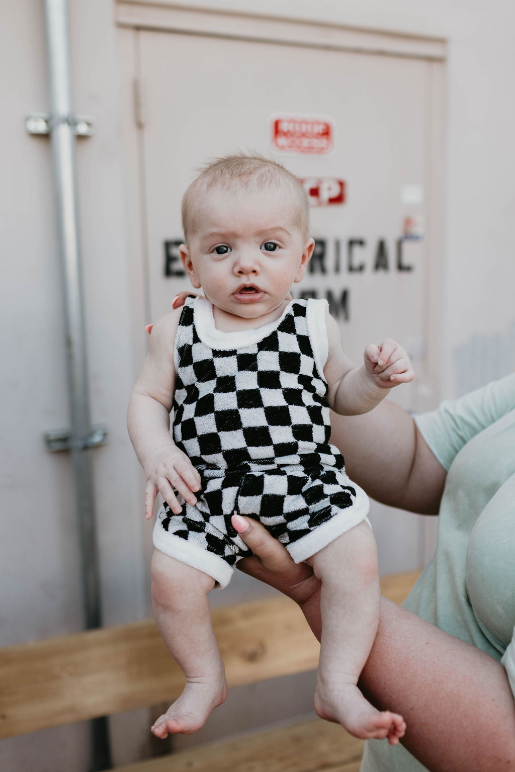 An adult is holding a curious-looking baby with light hair, dressed in the trendy Forever French Terry Tank Short Set in a stylish Black + White Checker print, in front of a door labeled "Electrical Room." The background showcases part of the door and includes a bench.