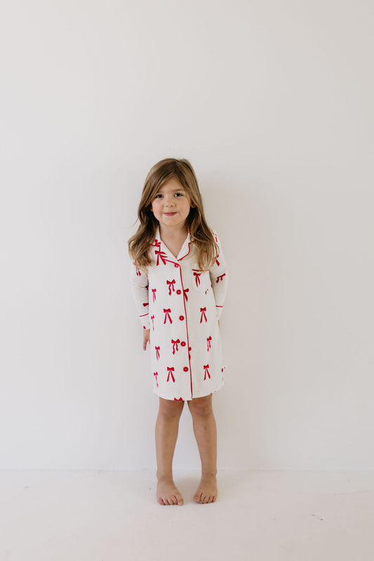 A young girl stands barefoot against a simple white backdrop, dressed in the Toddler Bamboo Sleeping Dress | Ribbons & Bows by forever french baby. This long-sleeved, knee-length dress features a soft bamboo fabric adorned with a red bow pattern. Her brown hair cascades gently around her shoulders, and she displays a calm expression.