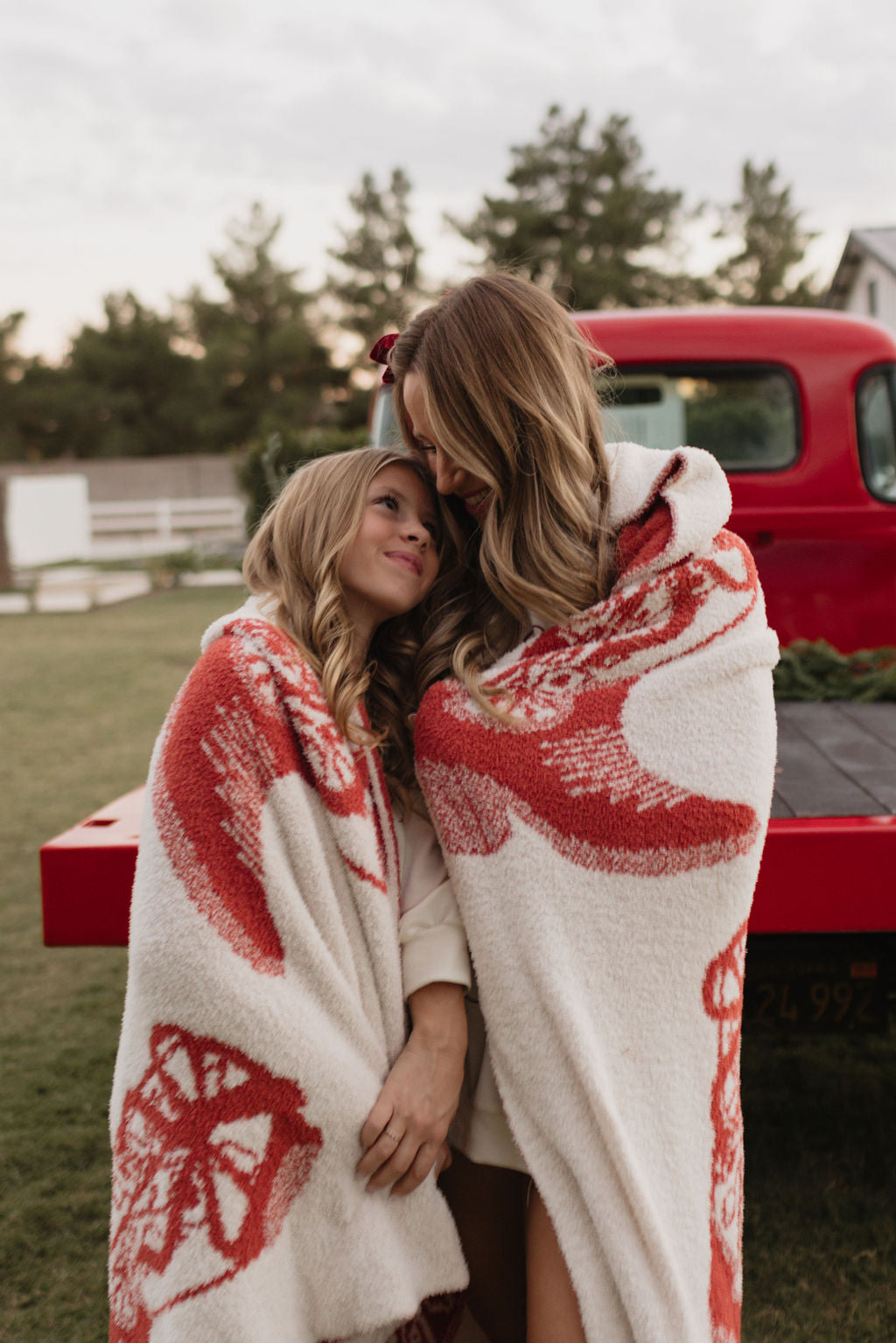 Two women wrapped in a lolo webb Plush Blanket | Cowgirl Christmas, standing beside a red vintage truck outdoors. The taller woman gently leans her head on the other's shoulder. Both with long hair and warm smiles, they cherish the serene moment together.