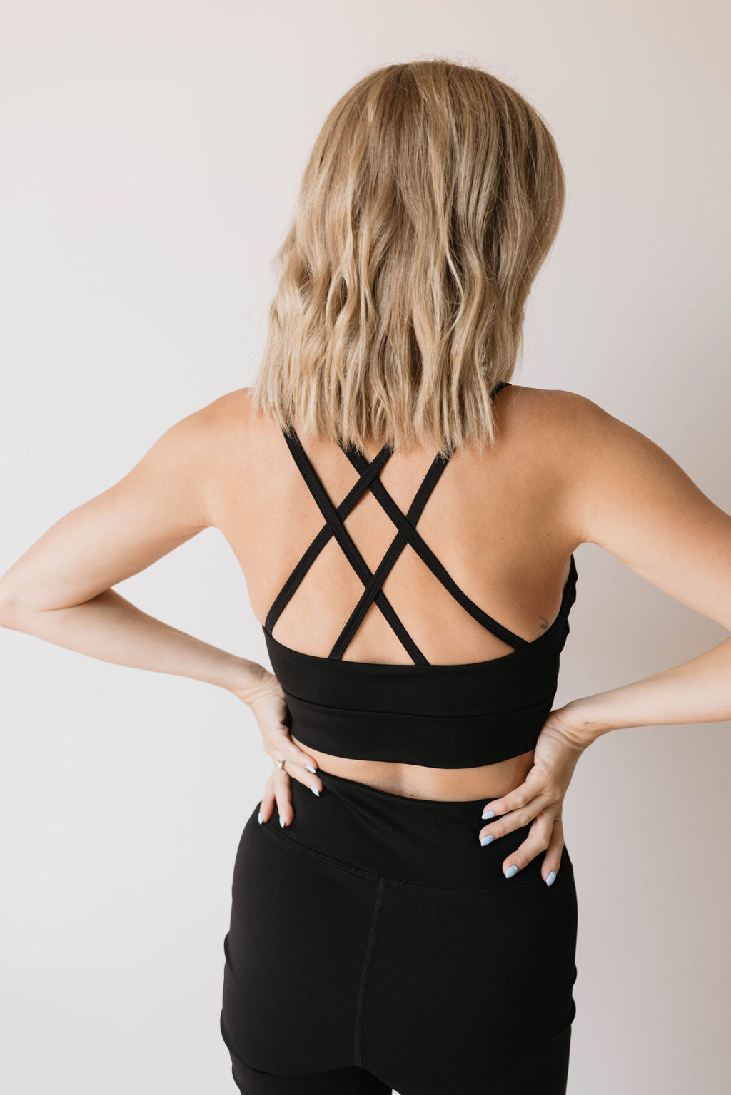 A woman with shoulder-length blonde hair stands with her back to the camera, confidently wearing the forever french baby Women's Workout Set in black. The sleek active wear ensemble includes an athletic top adorned with crisscross straps and matching leggings. Her hands rest assertively on her hips.