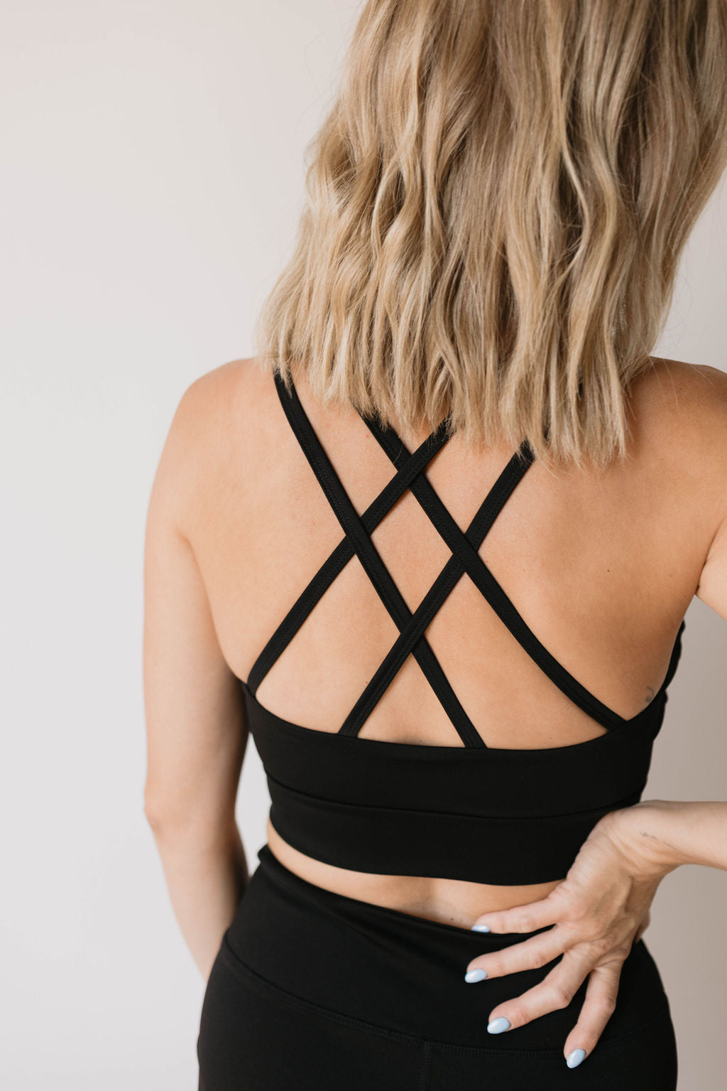 A woman with shoulder-length blonde hair is shown from the back, wearing the Women's Workout Set in Black from forever french baby, which features crossed straps and matching black leggings. Her right hand is placed on her hip, showcasing light blue painted nails.