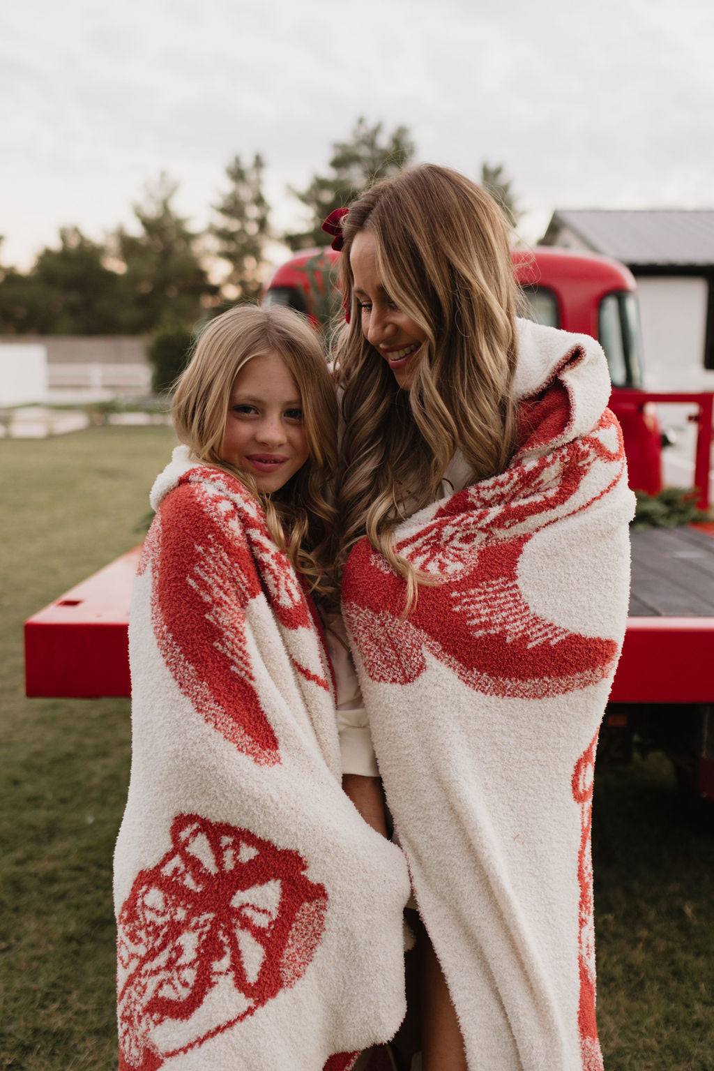 A woman and a girl are wrapped in the Plush Blanket from lolo webb's Cowgirl Christmas collection, enjoying the outdoor setting. They exchange warm smiles in their cozy surroundings. In the background, a red vehicle is nestled among the trees, adding to the scene's warmth and charm with its inviting presence.