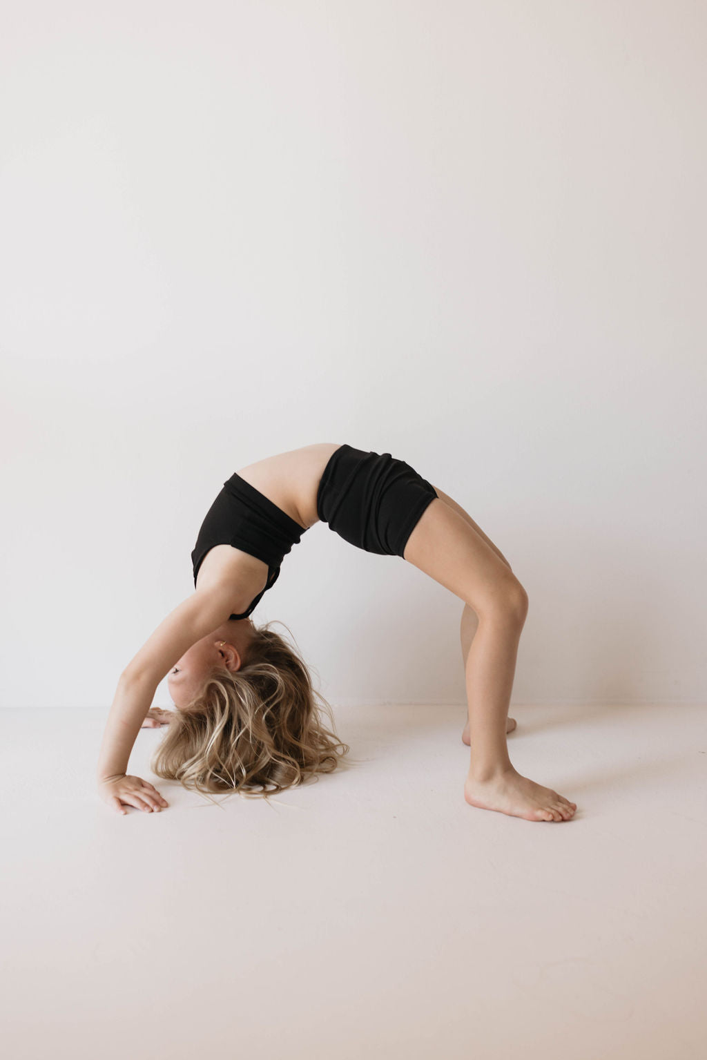 A young girl dressed in the Children's Workout Set in black from forever French baby performs an advanced backbend yoga pose, her hands and feet touching the ground with her back arched upwards. The background is a plain white wall.