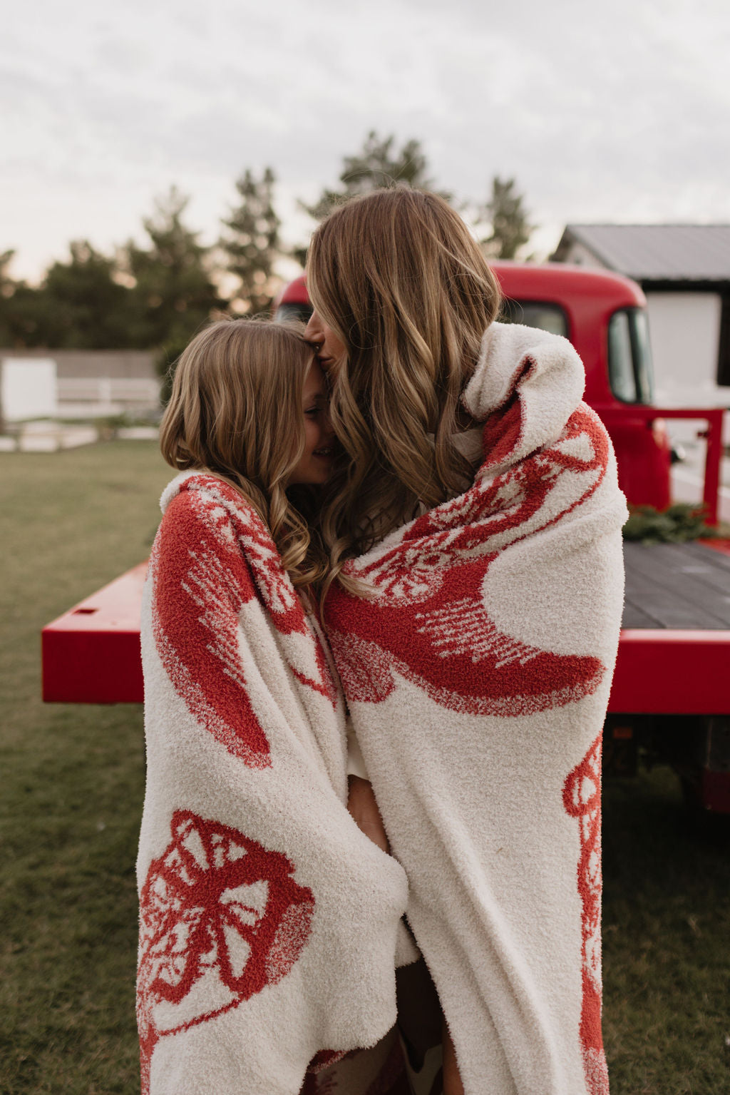 A woman and a child are wrapped in the dreamy softness of lolo webb's Cowgirl Christmas Plush Blanket, crafted from microfiber feather yarn. They stand close together outdoors, enjoying the cozy embrace amid a grassy field and trees in front of a red vehicle, confident in the safety and comfort of their Oeko-Tex certified blanket.