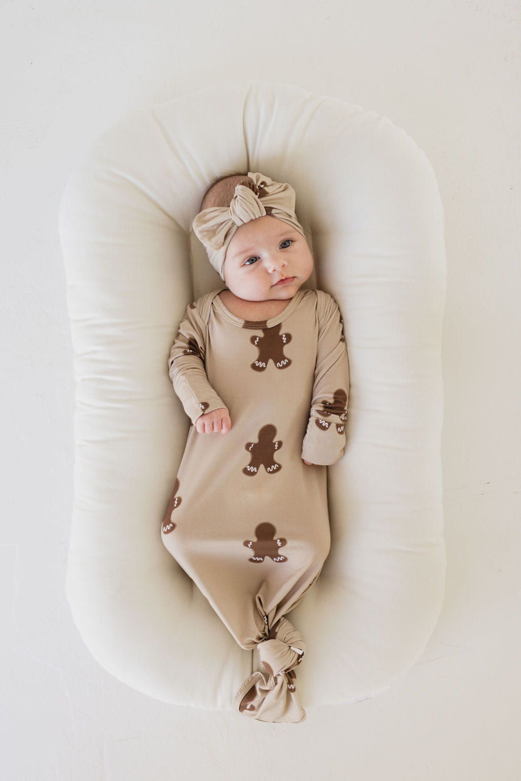 A baby lies on a soft, white cushion, wearing a beige outfit adorned with brown teddy bear designs and an adorable Gingerbread Bamboo Head Wrap from forever french baby. The baby's arms are slightly raised against a plain, light-colored background.