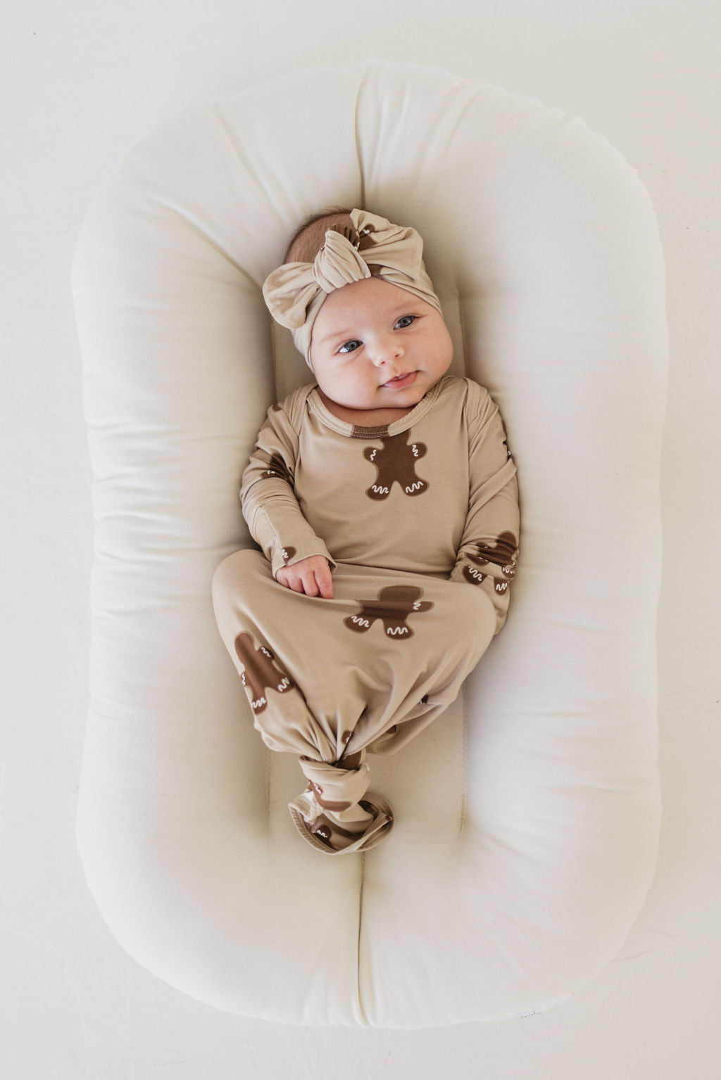 A baby is swaddled in a brown outfit with bear patterns, lying on a white cushioned surface. The baby is wearing the adorable "Bamboo Head Wrap | Gingerbread" by forever french baby, complete with a matching bow, and has a calm expression.