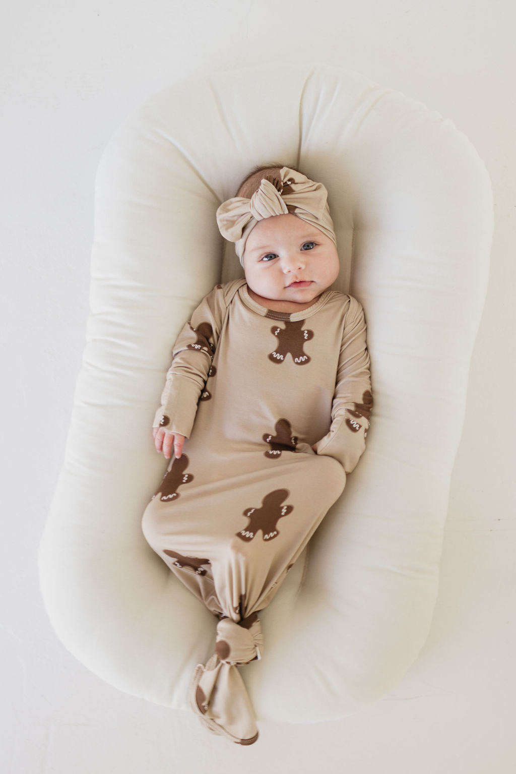 A baby wearing a printed beige onesie and a "Bamboo Head Wrap | Gingerbread" from forever french baby lies on a white cushioned surface, looking up.