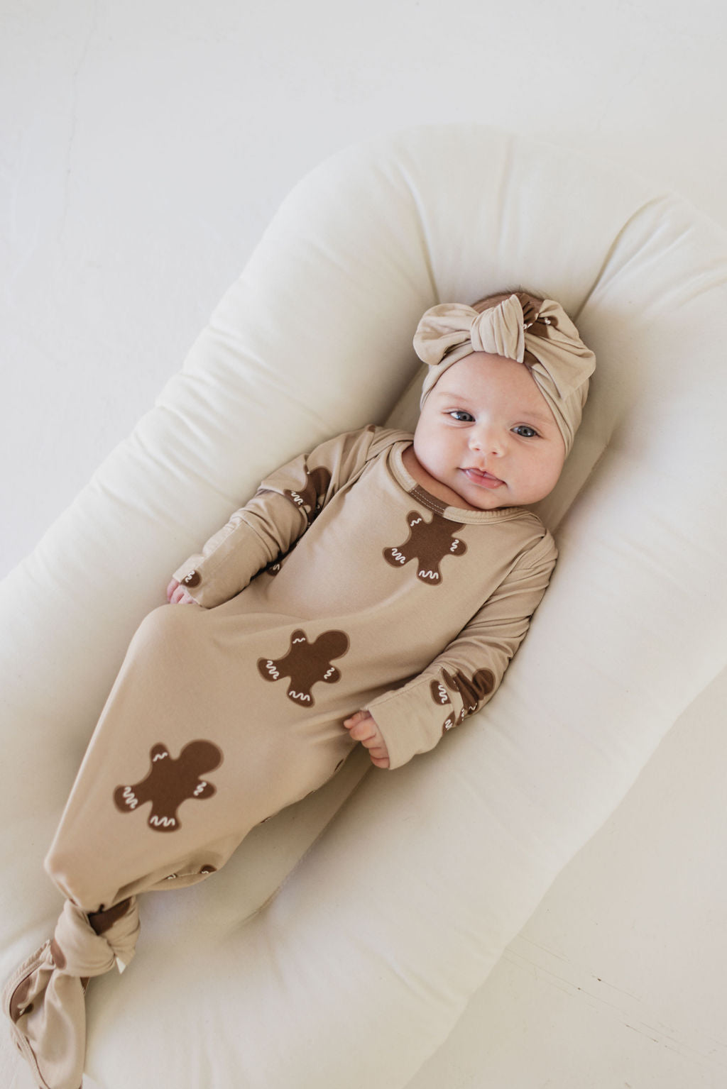 A baby is lying on a soft white pillow, wearing a light brown outfit adorned with gingerbread man patterns. The baby is also sporting the Bamboo Head Wrap in Gingerbread by forever french baby, which perfectly matches the attire, as they look calmly at the camera.