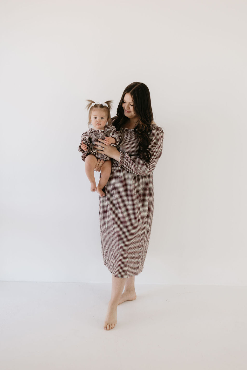 A woman with long hair holds a baby, both in matching chocolate gingham. She wears a checkered dress, and the baby is in a "Ruffle Romper | Chocolate Chai" by forever french baby. They stand barefoot against a plain white background.