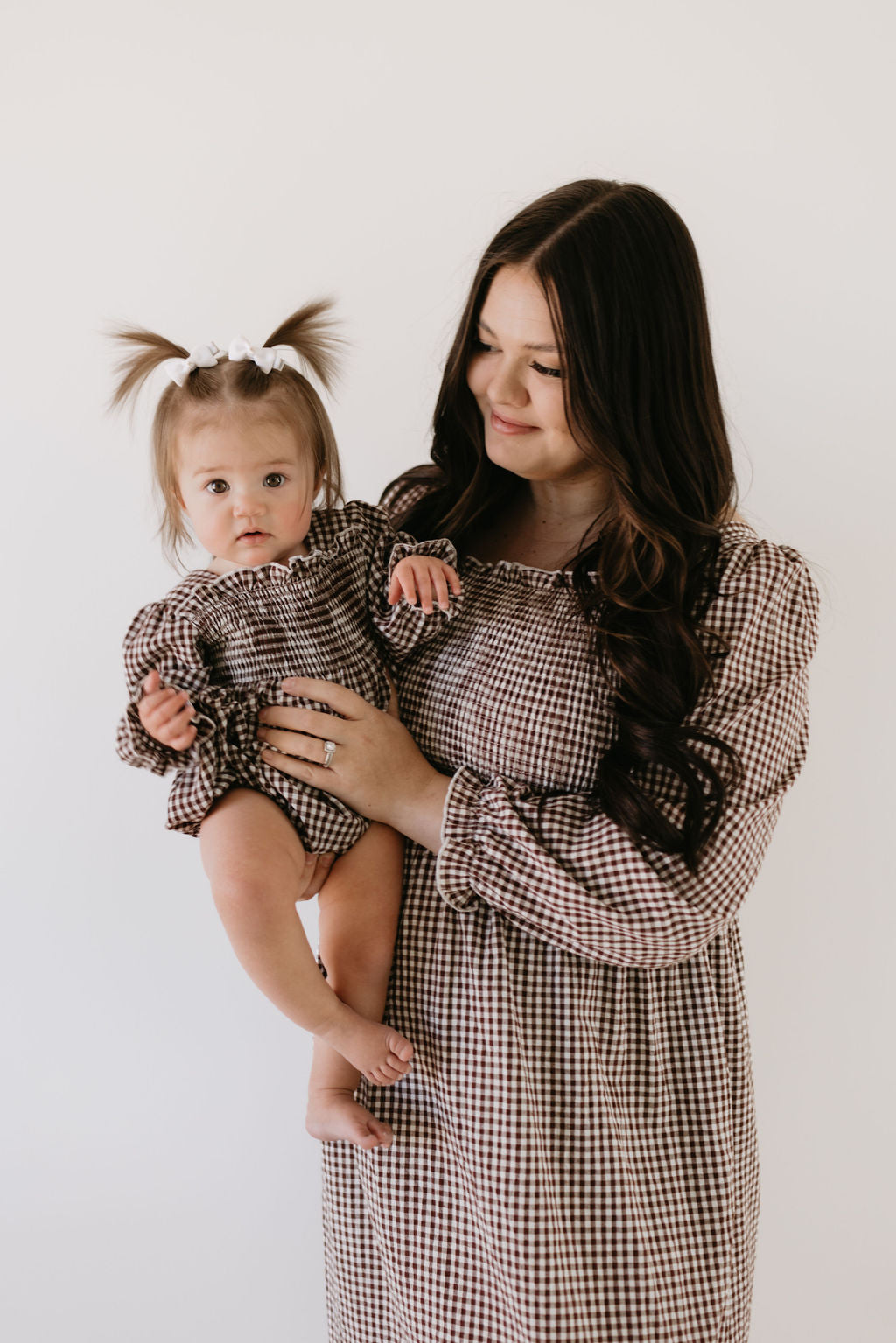 A woman with long dark hair holds a baby in pigtails, both wearing matching chocolate gingham dresses. The baby sports the "Ruffle Romper" by forever french baby. Their gentle smiles stand out against the plain white background.