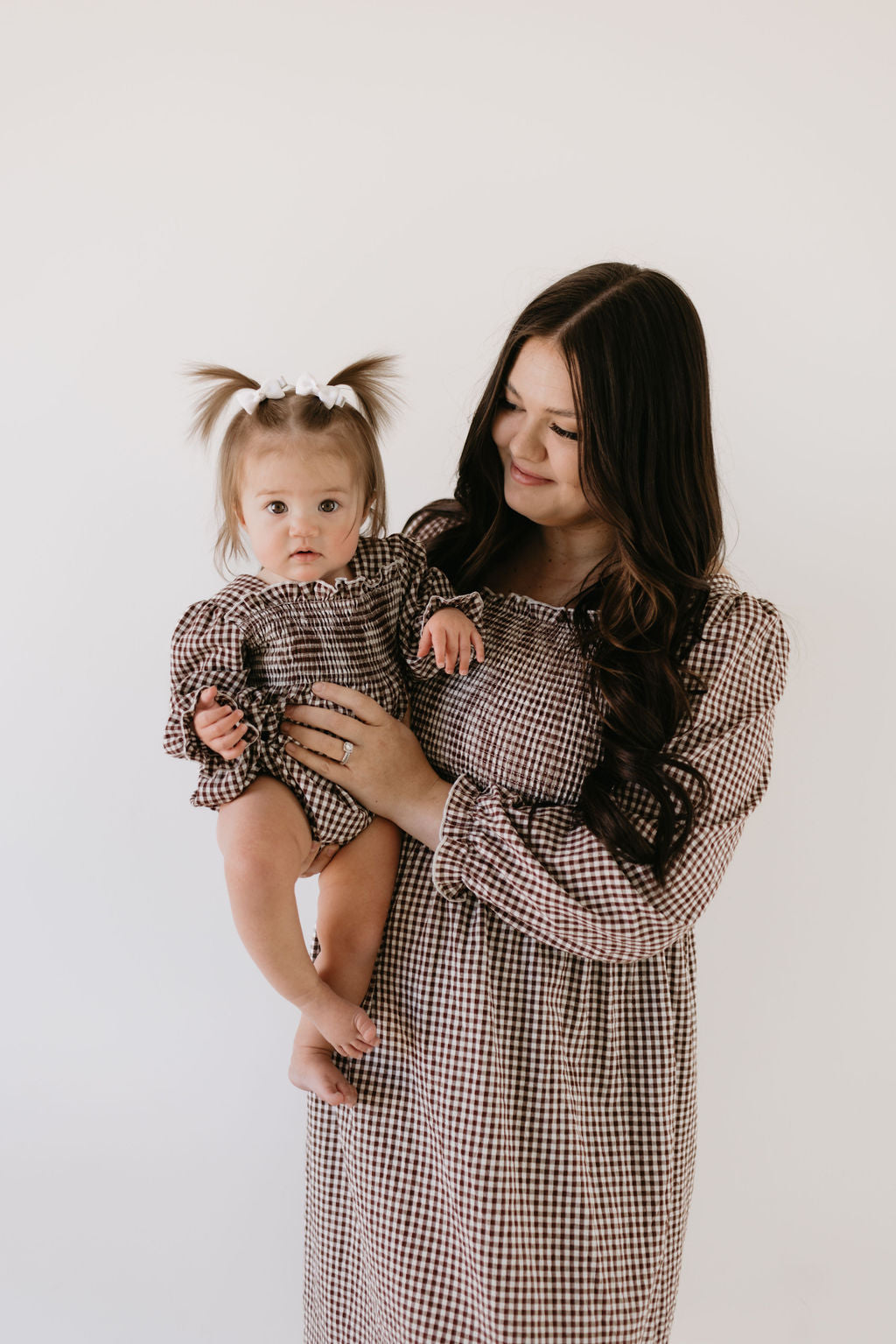 A woman with long dark hair in a chocolate gingham pattern dress holds a baby wearing the "Ruffle Romper | Chocolate Chai" by forever french baby. They both smile at the camera against a plain white background, their matching outfits creating an endearing, harmonious scene.