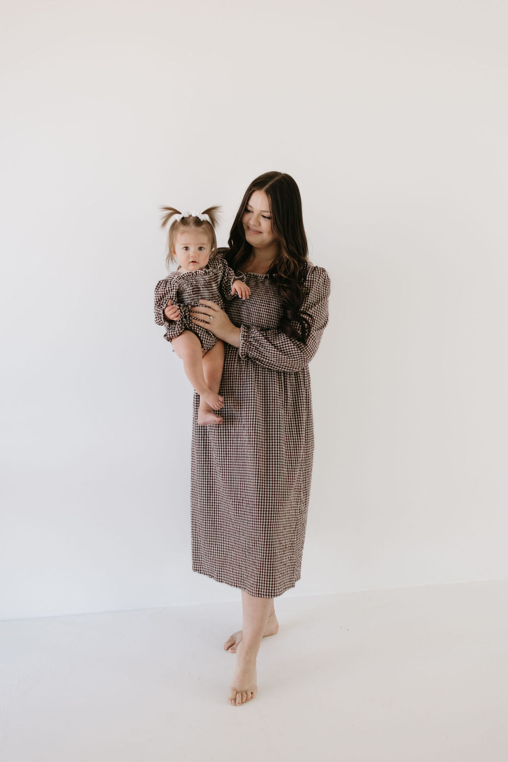 A dark-haired woman in a long checkered dress holds her baby, both wearing matching outfits. The baby, with two pigtails, wears the "Ruffle Romper | Chocolate Chai" by forever french baby. They are barefoot against a plain white background.