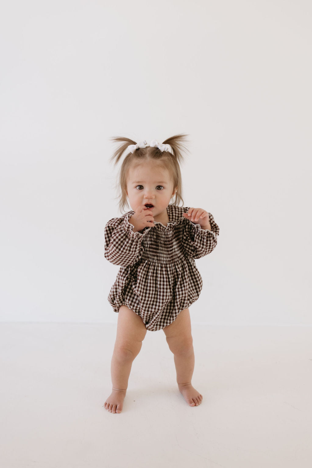 A toddler with two ponytails tied with white ribbons stands barefoot against a white background, wearing the forever french baby Ruffle Romper in Chocolate Chai gingham, with an expressive look on her face.