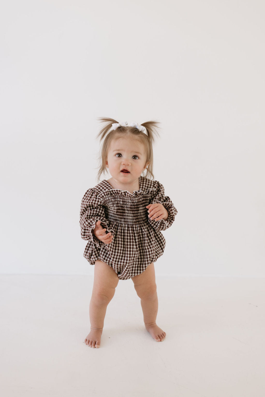 A toddler in a "Ruffle Romper | Chocolate Chai" by forever french baby stands barefoot against a light background. The child sports small ponytails and looks forward with one hand slightly raised.