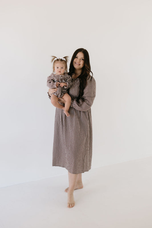 A woman with long hair holds a baby, both smiling against a plain background. They wear matching Chocolate Chai dresses from forever french baby, and the baby's two small ponytails add to their charm.