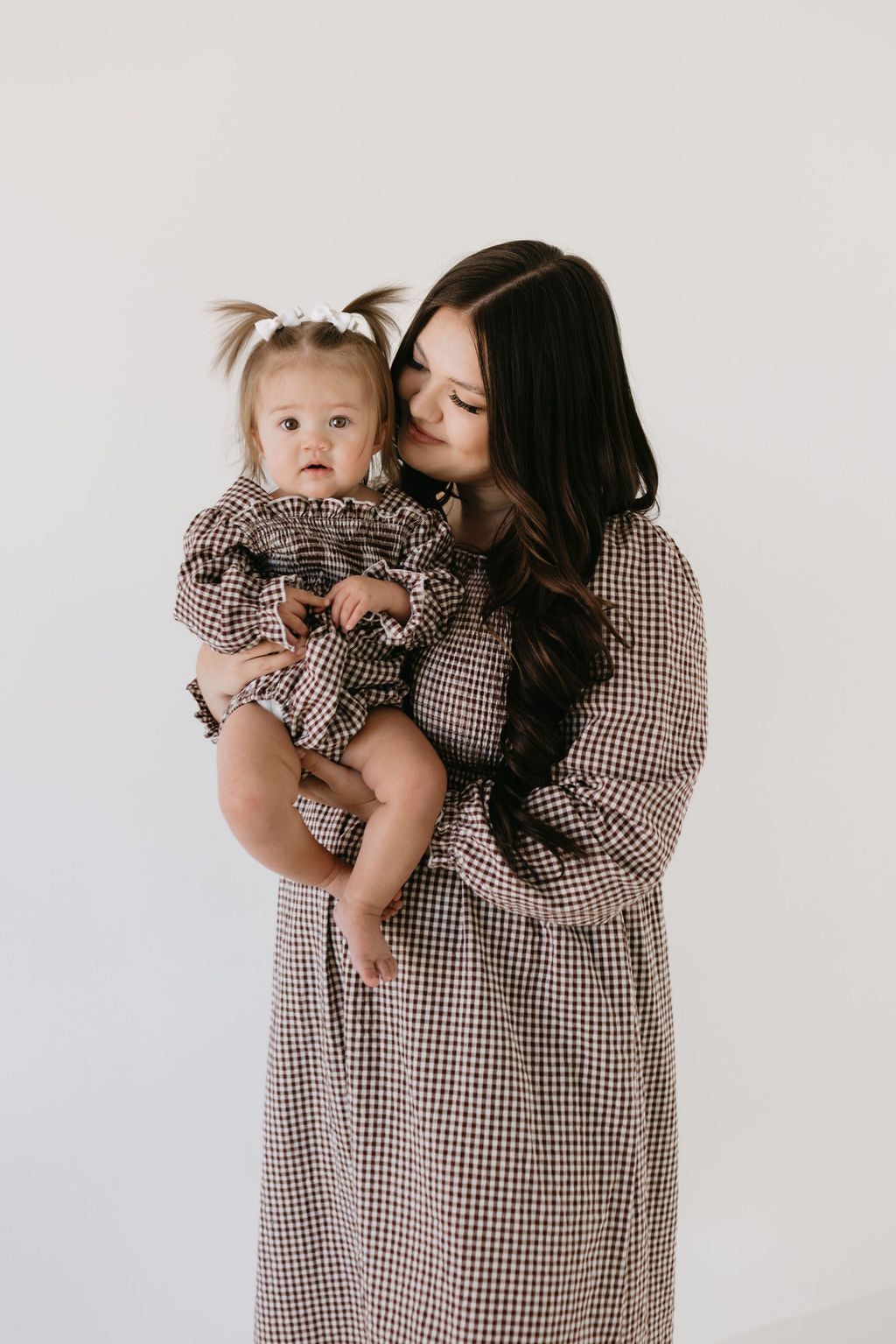 A woman with long dark hair wears a forever french baby Women's Long Sleeve Dress in Chocolate Chai while holding a baby in matching attire. The baby has pigtails adorned with a white bow. Against a plain white background, the woman gazes lovingly at the child.