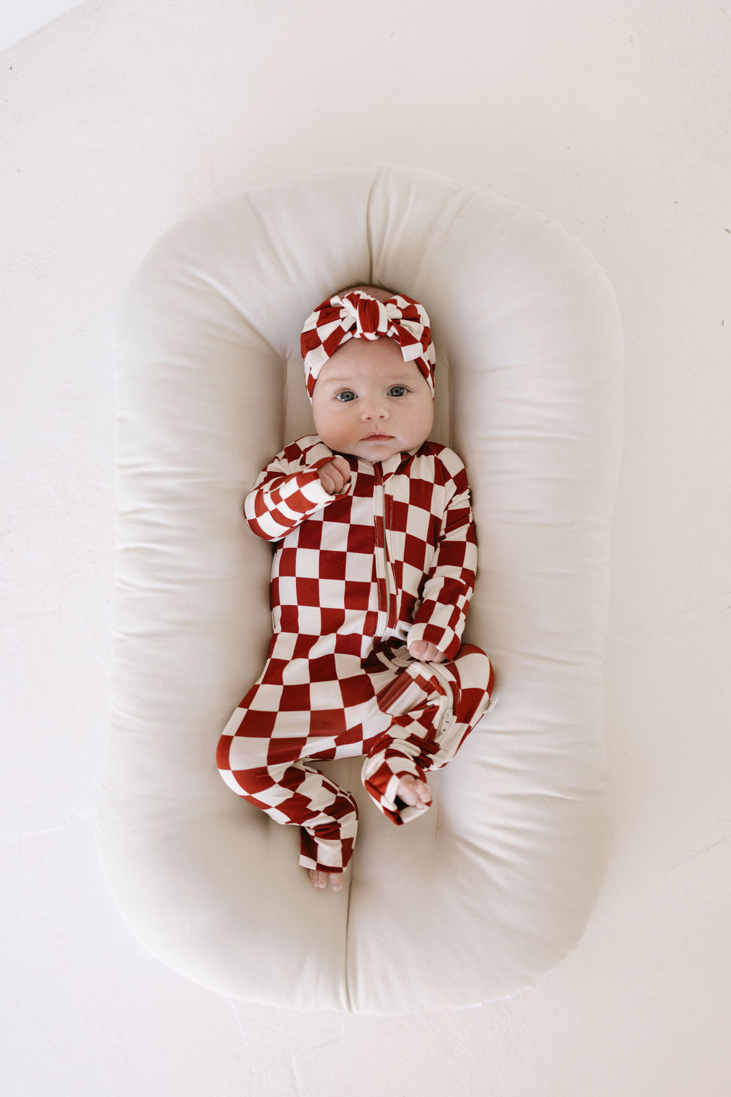 A baby lies on a white cushioned surface, wearing a red and white checkered outfit with the Bamboo Head Wrap by lolo webb, known as the Quinn. The baby's eyes are open, gazing upward with curiosity.