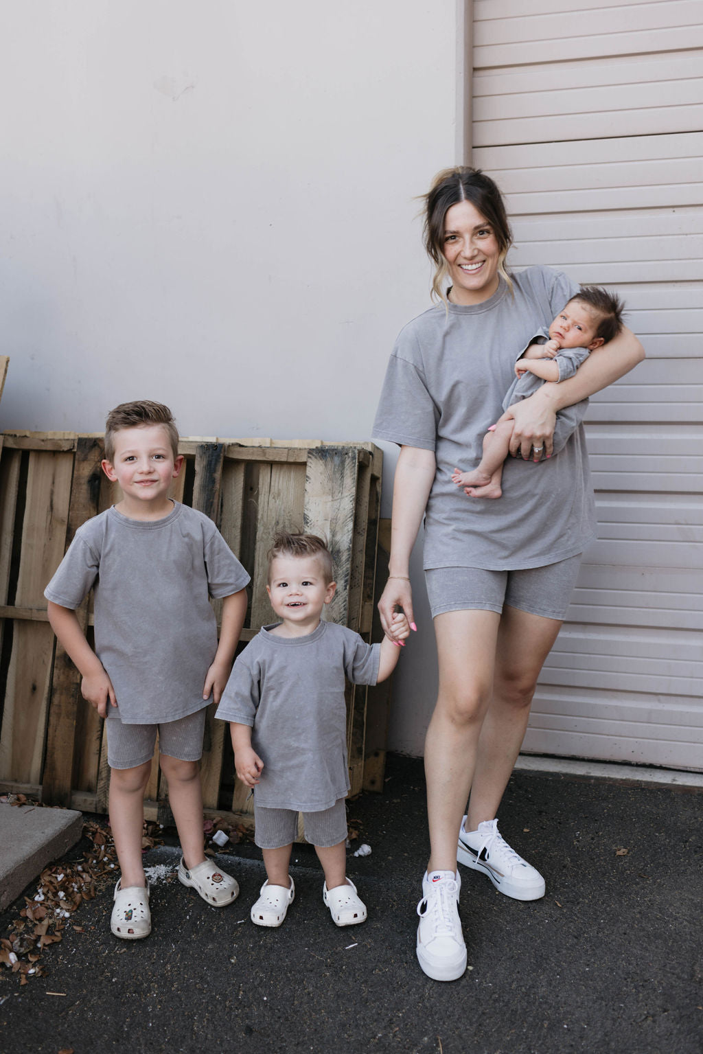 A woman stands next to a garage door holding a baby, with two young boys standing beside her. All four individuals are dressed in matching grey outfits made of organic cotton. The boys wear the Vintage Washed Steel Children's Short Sets from Forever French Baby, while the woman wears grey shorts and a t-shirt, holding the baby in a Forever French Baby outfit.