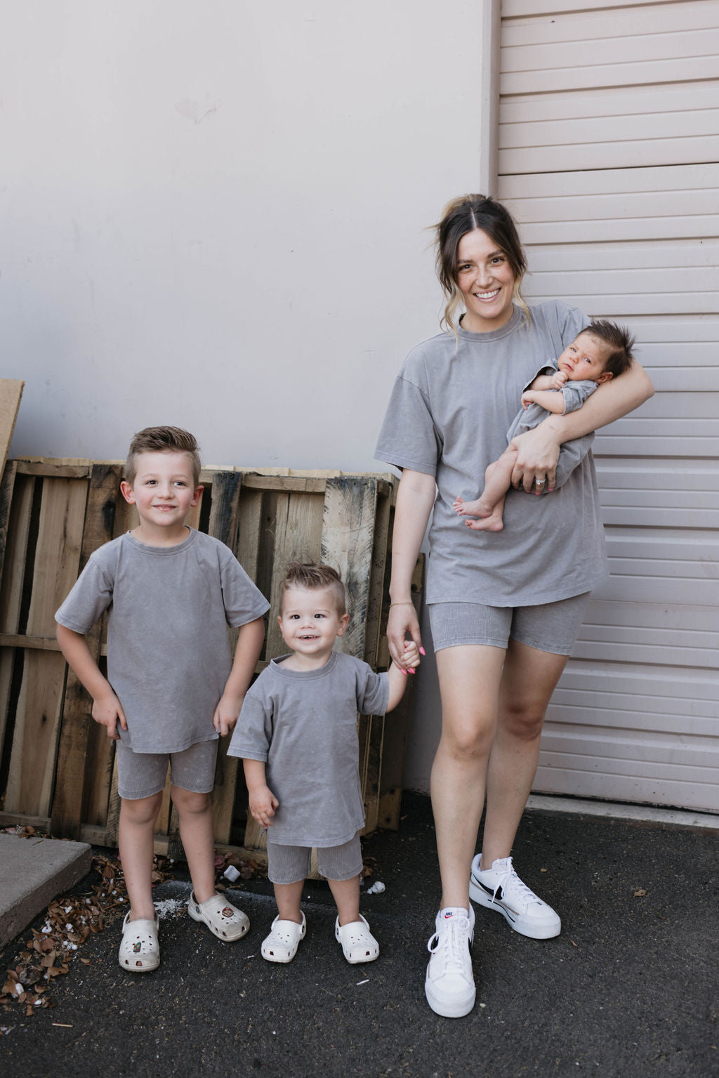 A woman stands in front of a garage door, holding a baby in her right arm and the hand of a toddler with her left. Another young child stands beside them. They all sport matching grey outfits, including stylish ribbed biker shorts from the Women's Short Set | Vintage Washed Steel collection by forever french baby. Crates are stacked against the wall behind them.