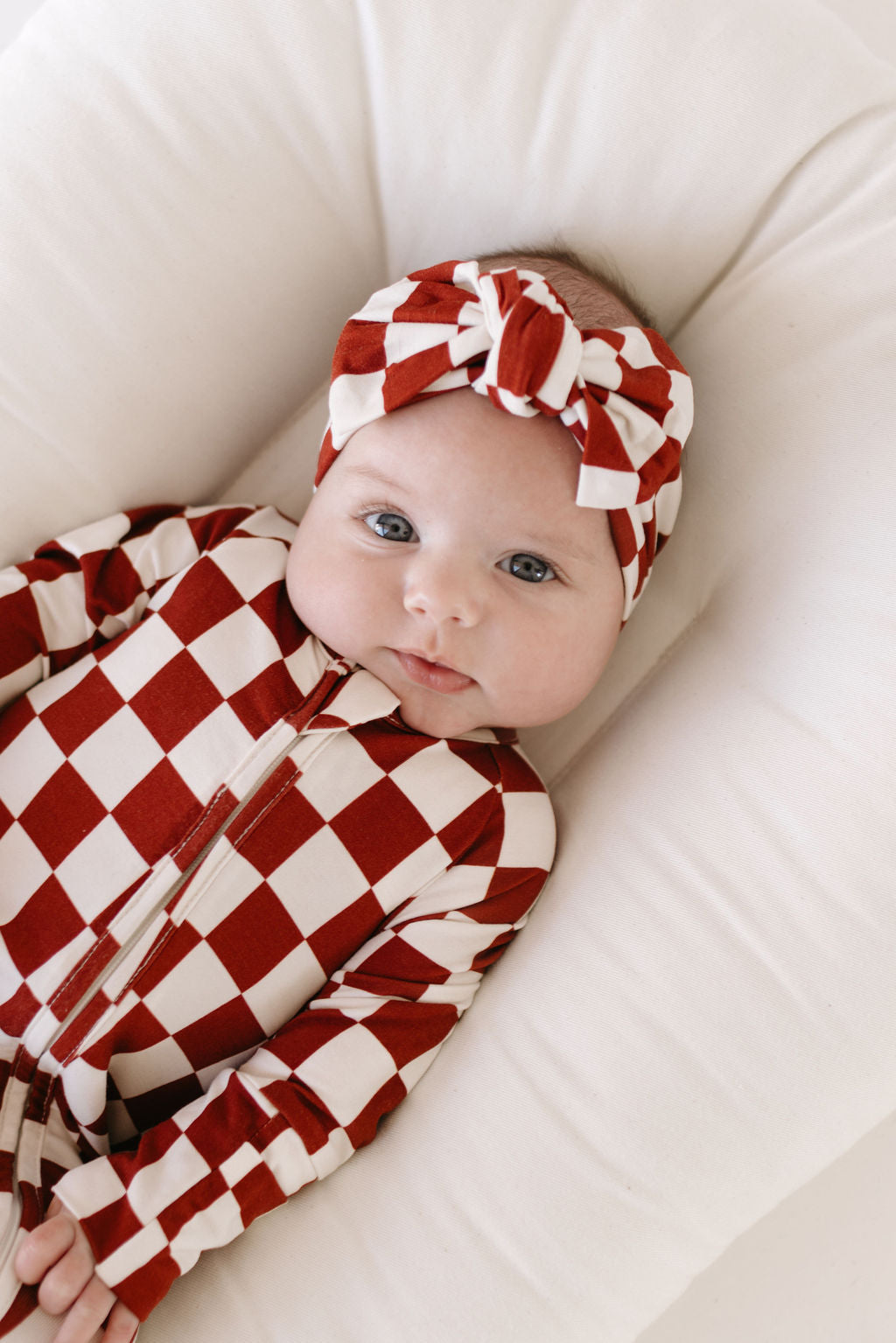 A baby lies on a white cushion, wearing a red and white checkered outfit, complemented by "the Quinn" Bamboo Head Wrap by lolo webb. The baby's bright eyes are gazing directly at the camera.