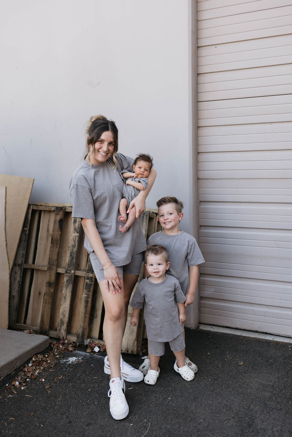 A woman stands outside in front of a garage door, holding a baby dressed in an adorable outfit from Forever French Baby. Two young boys, wearing Forever French Baby's Vintage Washed Steel Children's Short Sets made of organic cotton, stand next to her. They all wear matching gray outfits. A wooden pallet leans against the wall behind them as they smile warmly.