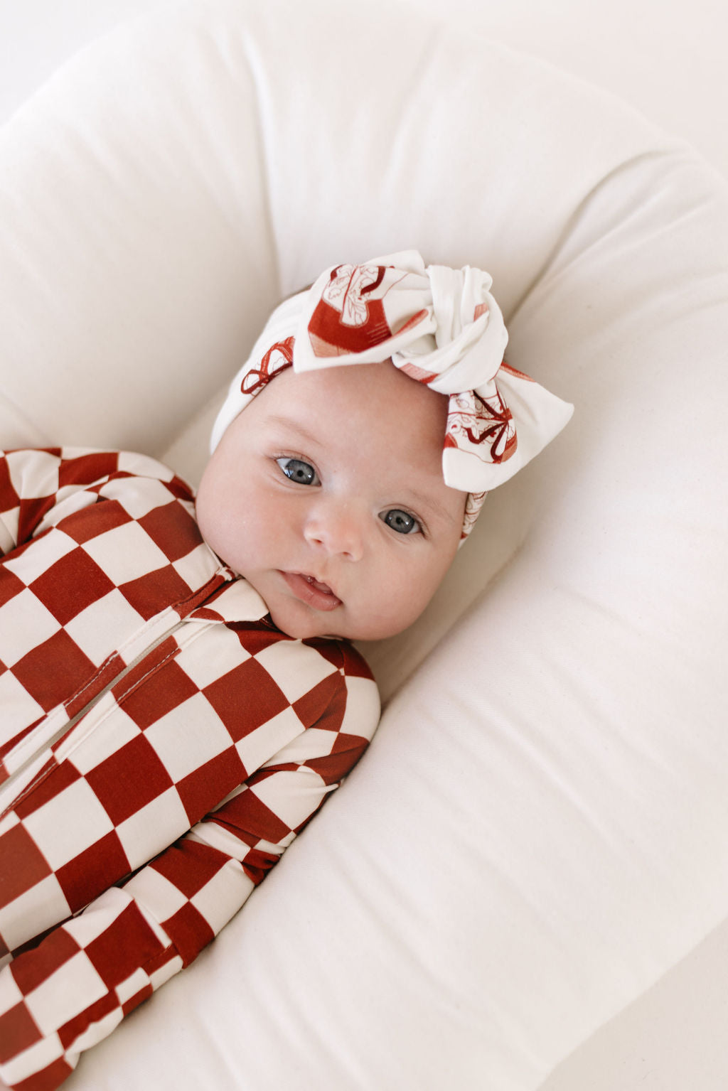 A baby dressed in the Cowgirl Christmas outfit, featuring a red and white checkered design with a matching adjustable Bamboo Head Wrap from lolo webb, lies on a white cushion, gazing up with a calm expression.