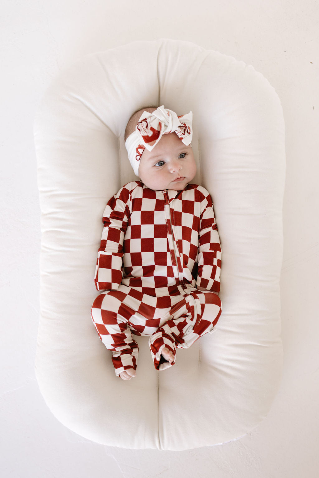 A baby lies on a soft white cushion, wearing a red and white checkered onesie and the Lolo Webb "Bamboo Head Wrap | Cowgirl Christmas," adorned with an adjustable bow. The baby gazes upward, hands resting by their sides.