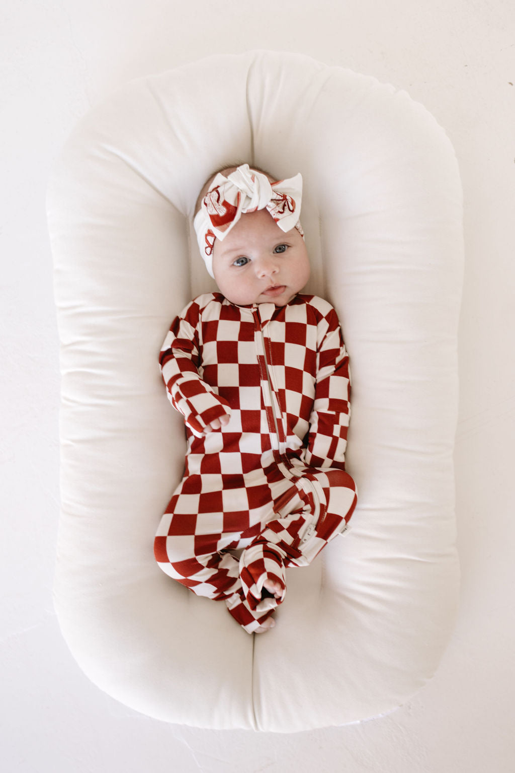 A baby lying on a white cushion wears the Cowgirl Christmas onesie with an adorable matching bamboo head wrap from lolo webb. The baby gazes upward with wide eyes.