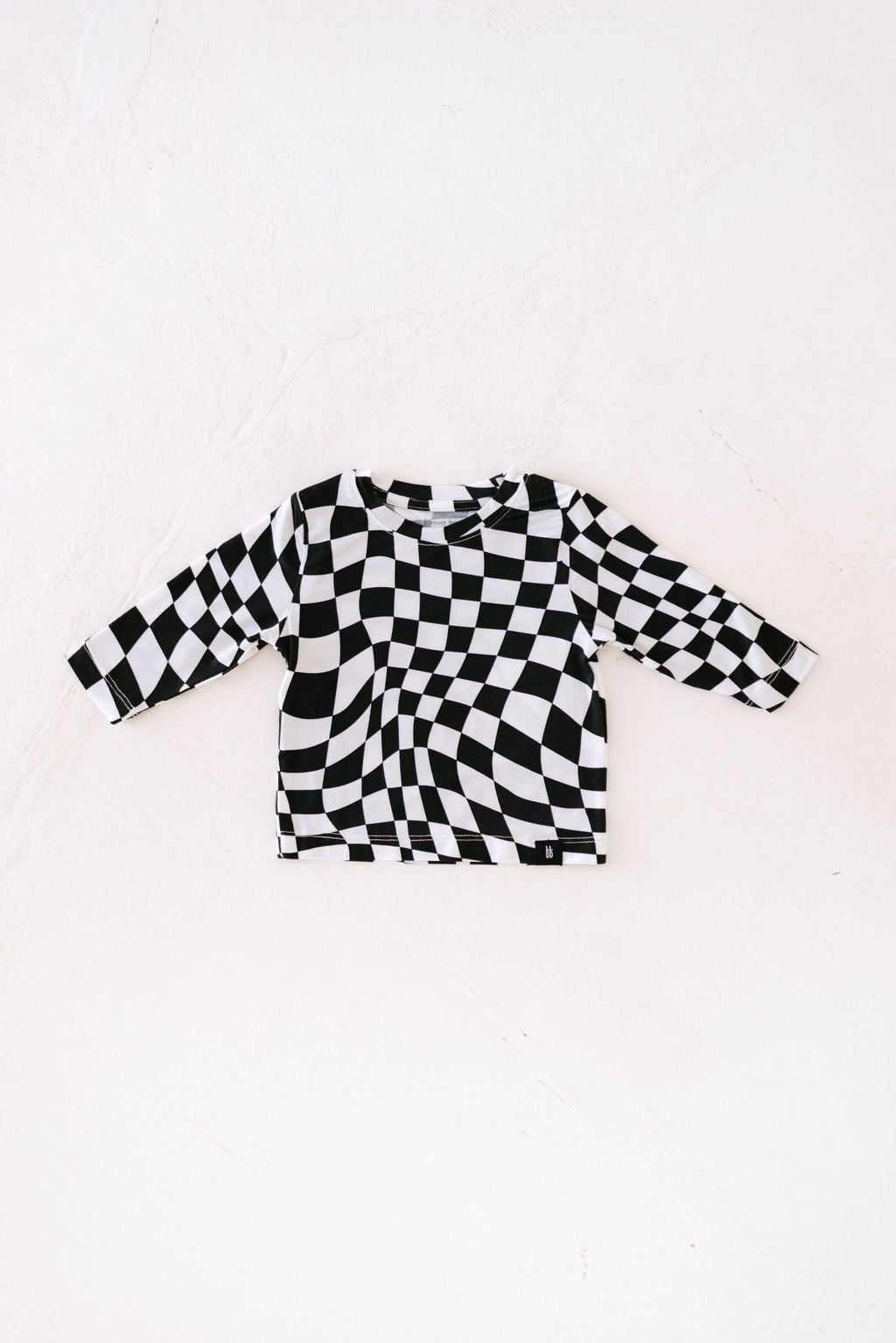 The Long Sleeve Bamboo T-Shirt by forever french baby, in a black and white wavy checkerboard pattern, is laid flat on a white background. Crafted from breathable bamboo fabric, its design creates an optical illusion effect.