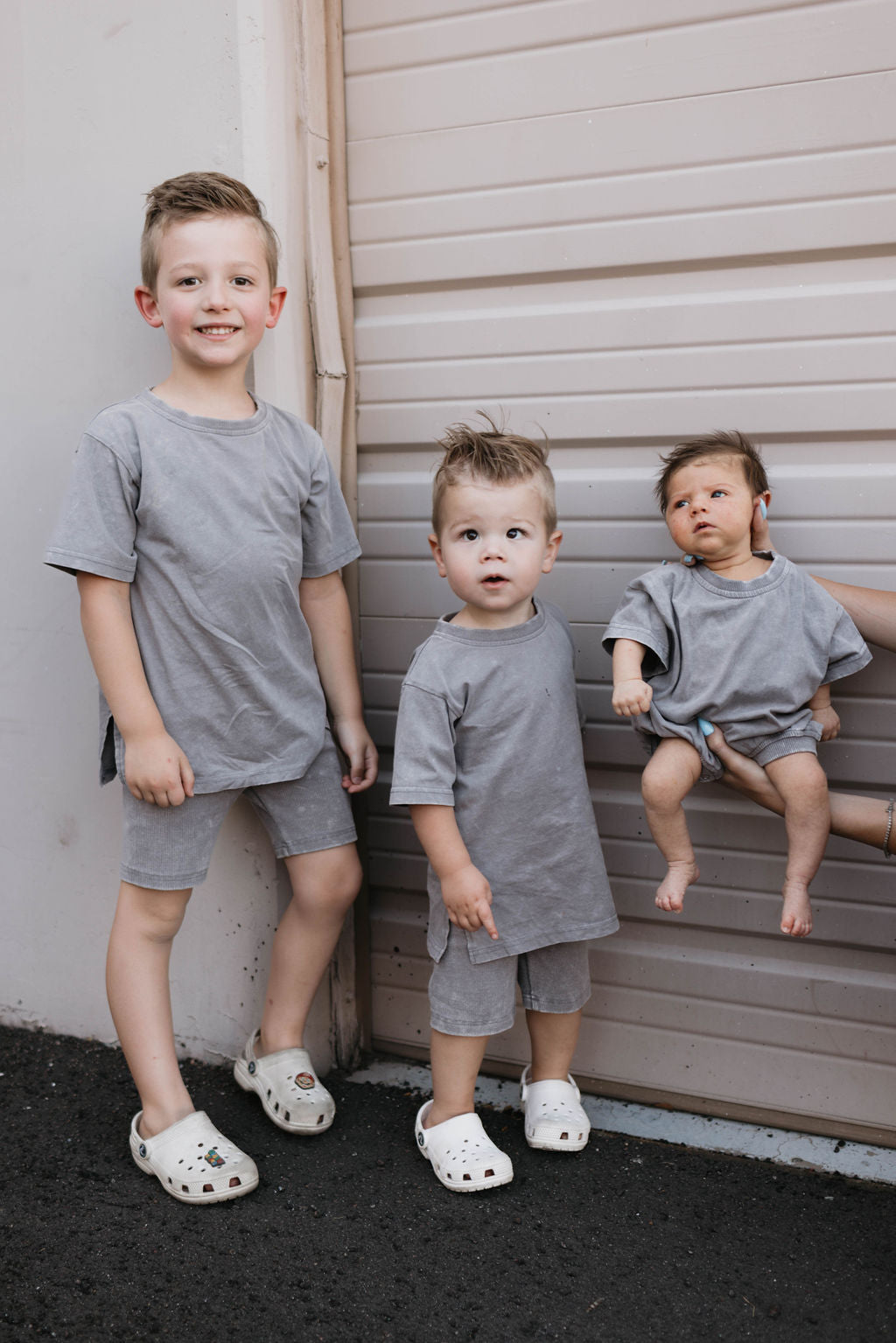 Three young children stand in front of a garage door. The older two boys stand on the ground, both wearing matching grey shirts, shorts, and white sandals. The youngest child is held by a hand in a Women's Short Set from forever french baby, specifically the Vintage Washed Steel design, also wearing a grey shirt and making a surprised expression.