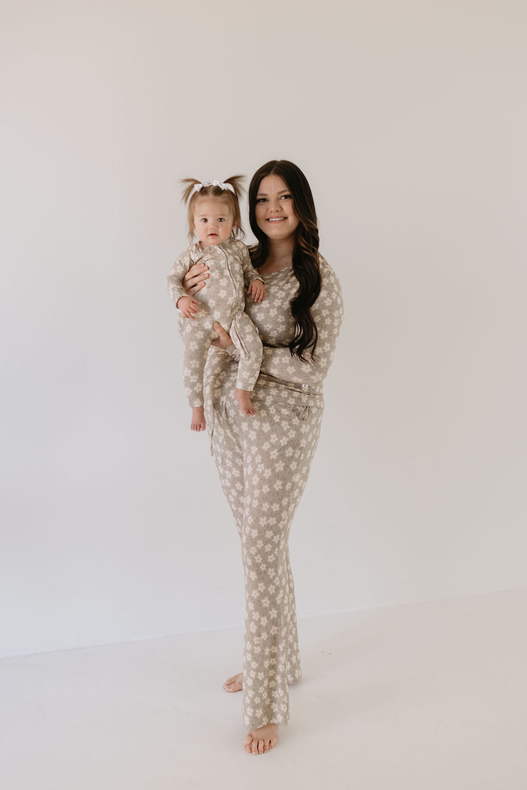 A woman and a toddler wearing the Bamboo Zip Pajamas in French Gray Floral from Forever French Baby stand against a plain background. The matching cozy outfits enhance their joyful expressions as the woman holds the pigtail-adorned toddler. Both are barefoot, wearing subtle smiles.