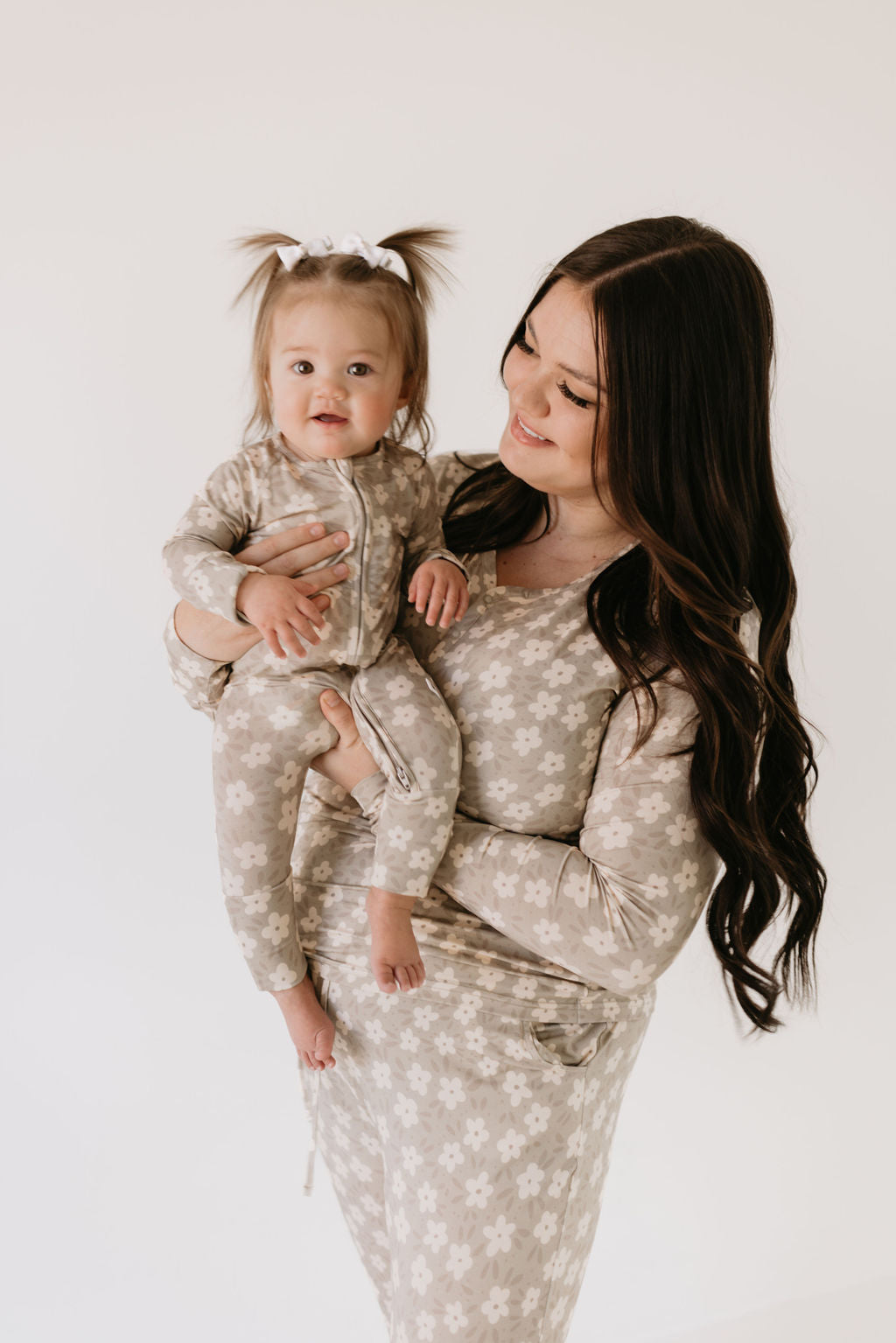 A woman with long dark hair is smiling while holding a toddler, both wearing Forever French Baby's Bamboo Zip Pajamas in French Gray Floral. The toddler's hair is styled in two small pigtails with a white bow, set against a plain white background.