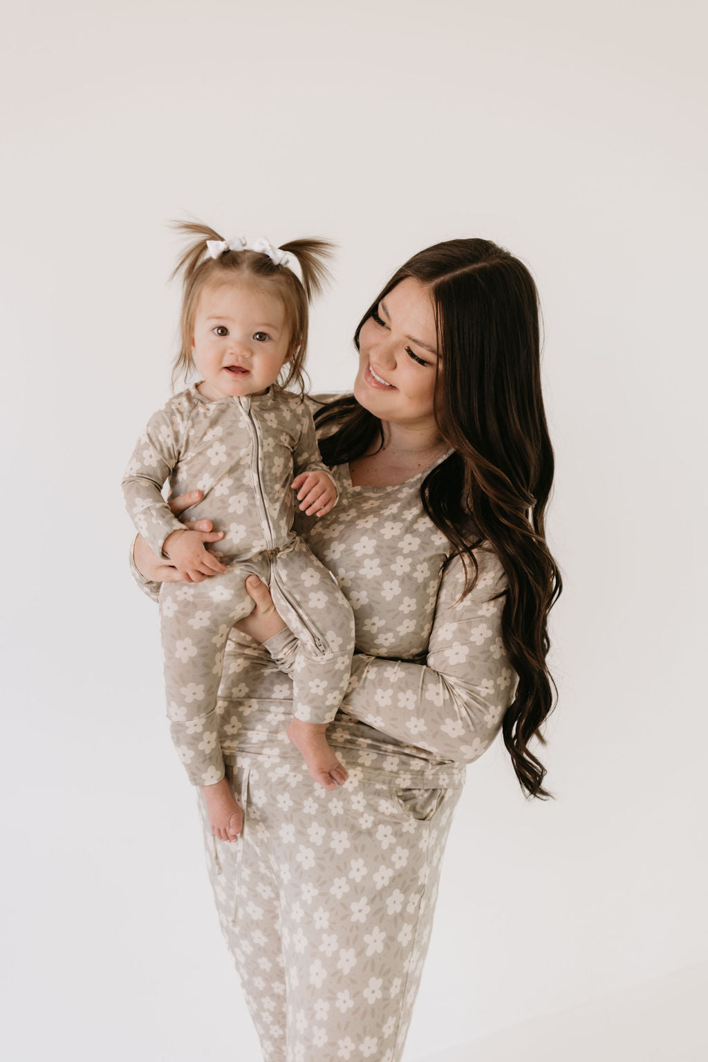 A woman with long brown hair, dressed in a floral-patterned outfit, holds a baby girl with pigtails who is wearing matching Bamboo Zip Pajamas in French Gray Floral by forever french baby. Both are smiling and gazing at each other against a plain white background, exuding warmth and togetherness.