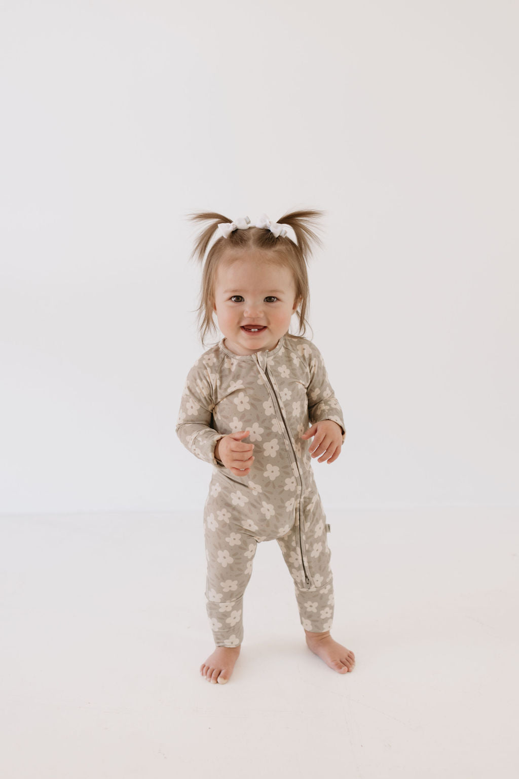 A toddler dressed in forever french baby's Bamboo Zip Pajamas in French Gray Floral stands barefoot with a white bow in her hair. She smiles at the camera with her arms slightly raised against the plain white background, beautifully showcasing this cozy sleepwear.