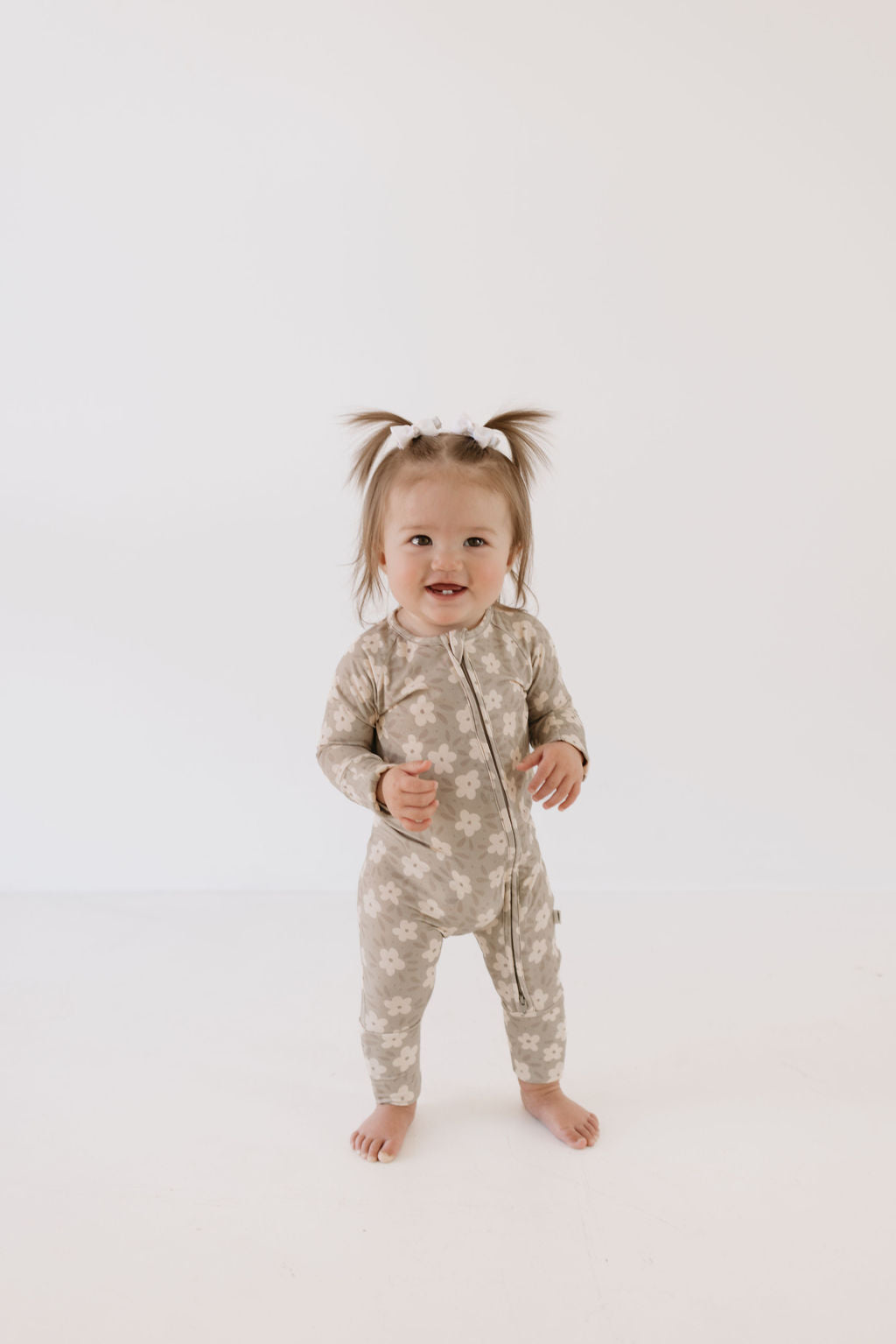 A toddler with short pigtails stands on a white background, wearing Forever French Baby's cozy Bamboo Zip Pajamas in a French Gray Floral pattern. These luxurious sleepwear pajamas ensure comfort and style as the child looks forward with a smile and bare feet.