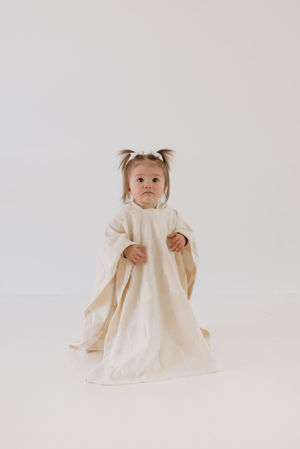 A toddler with two small pigtails wrapped in the cream-colored Hooded Towel | Cream Checkerboard from forever french baby stands against a plain white background, looking directly at the camera with a curious expression.