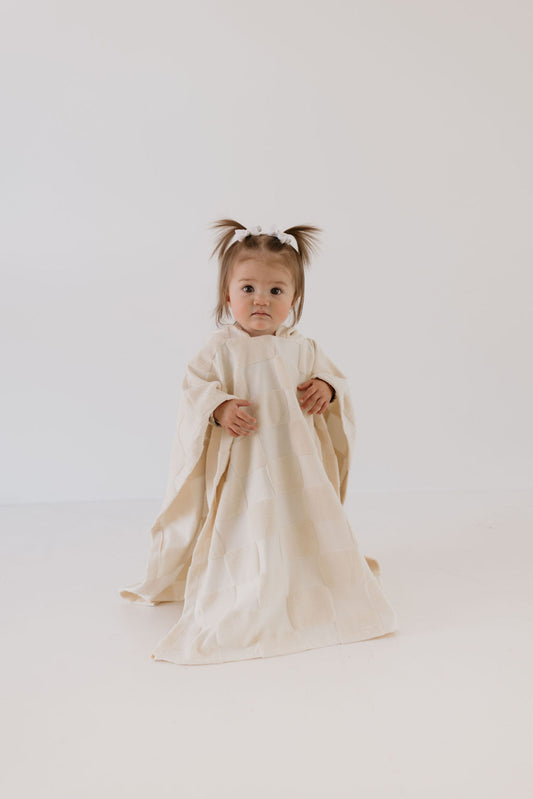 A toddler with two small pigtails stands on a white background, wrapped in an oversized robe from forever french baby. The Hooded Towel in Cream Checkerboard adds charm to the neutral expression, while its long, flowing sleeves evoke the cozy comfort of bath towels.