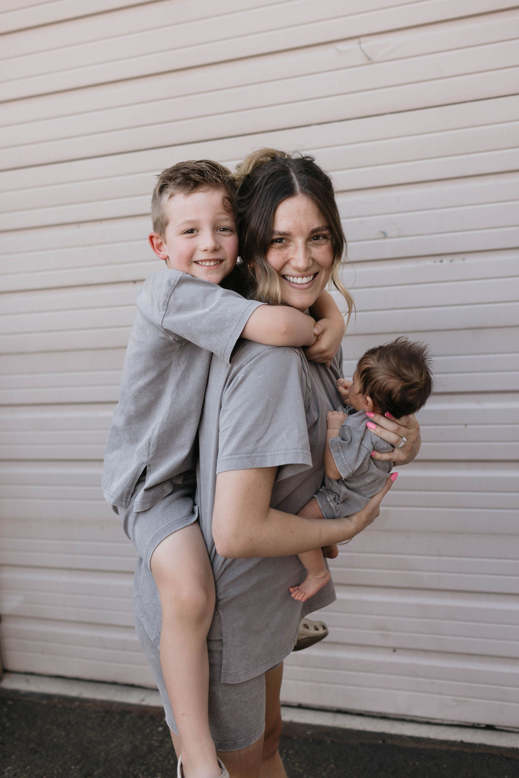 A woman smiles as she holds a young boy on her back and a baby in her arms, all dressed in matching gray outfits. Her ribbed biker shorts from the Women's Short Set | Vintage Washed Steel by forever french baby, combined with the kids' vintage washed steel tops, create a coordinated look against the beige garage door backdrop.