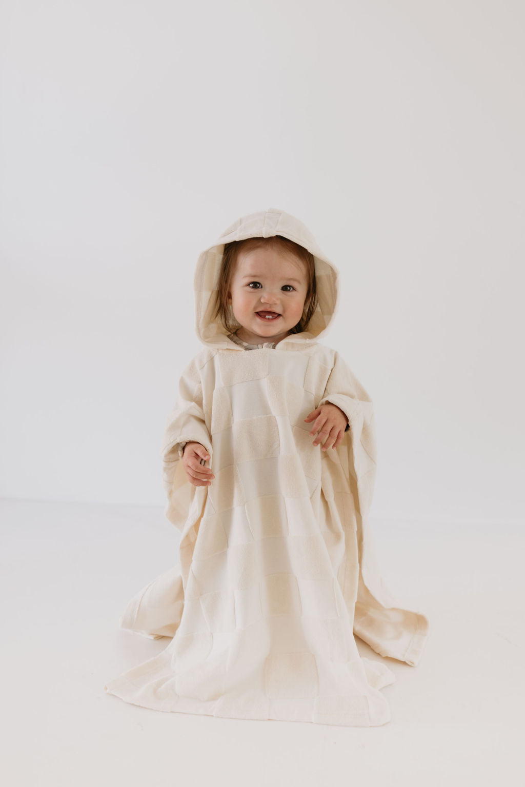 A cheerful toddler wearing a Hooded Towel in Cream Checkerboard from forever french baby stands against a plain white background. The oversized towel, reminiscent of soft bath towels, features a charming textured fabric, and the child's expression is playful.