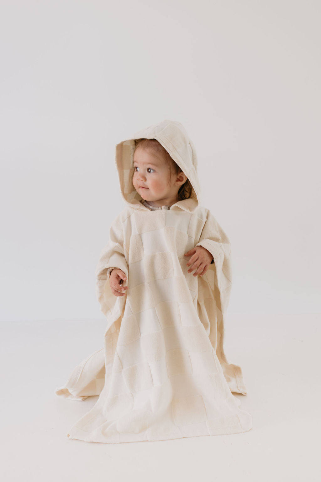 A baby sits on a white surface, enveloped in the Hooded Towel | Cream Checkerboard from forever french baby. The towel's subtle texture beautifully complements the baby's curious gaze, with a plain and minimalistic backdrop highlighting the brand's elegance.