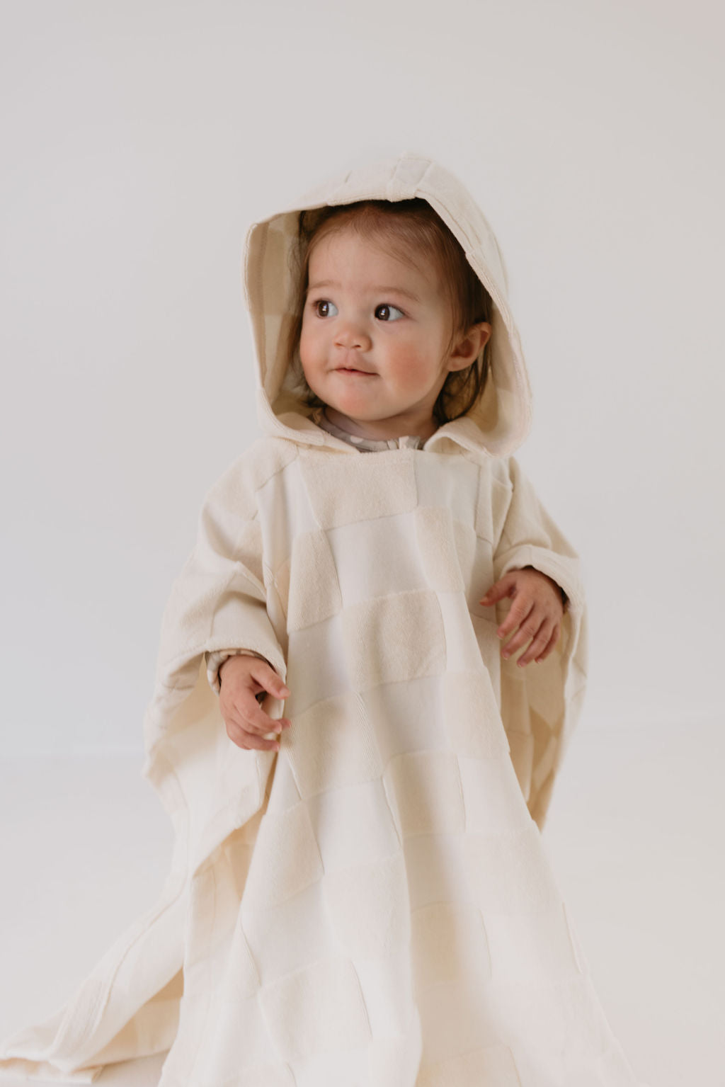 A baby wrapped in a Forever French Baby Hooded Towel in Cream Checkerboard looks to the side, smiling slightly. The towel drapes over their body, creating a cozy and adorable appearance against a neutral background—an image captured for the brand forever french baby.